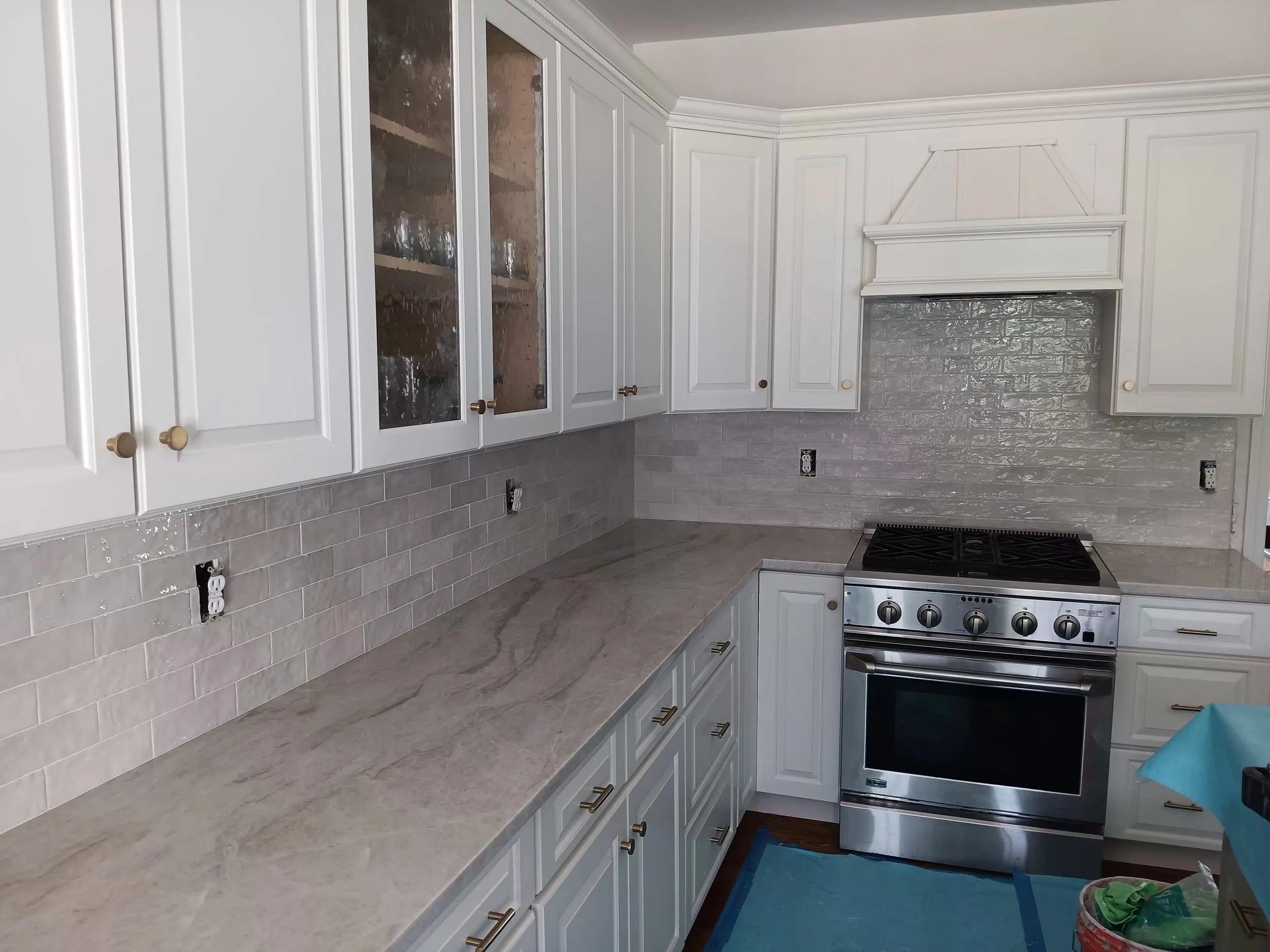 A kitchen under construction with white cabinets, a grey countertop, and a grey tiled backsplash. 