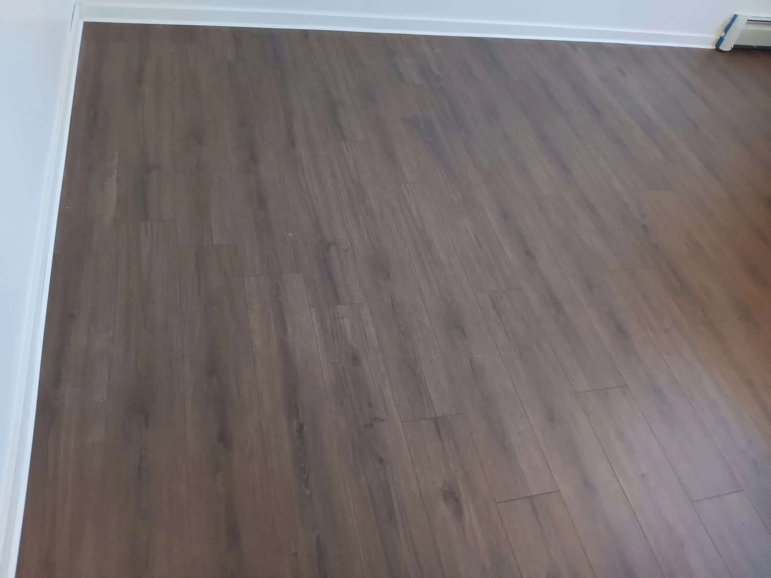 A room with dark brown wooden flooring and white walls with a white baseboard. There is a vent visible on the right side of the wall.
