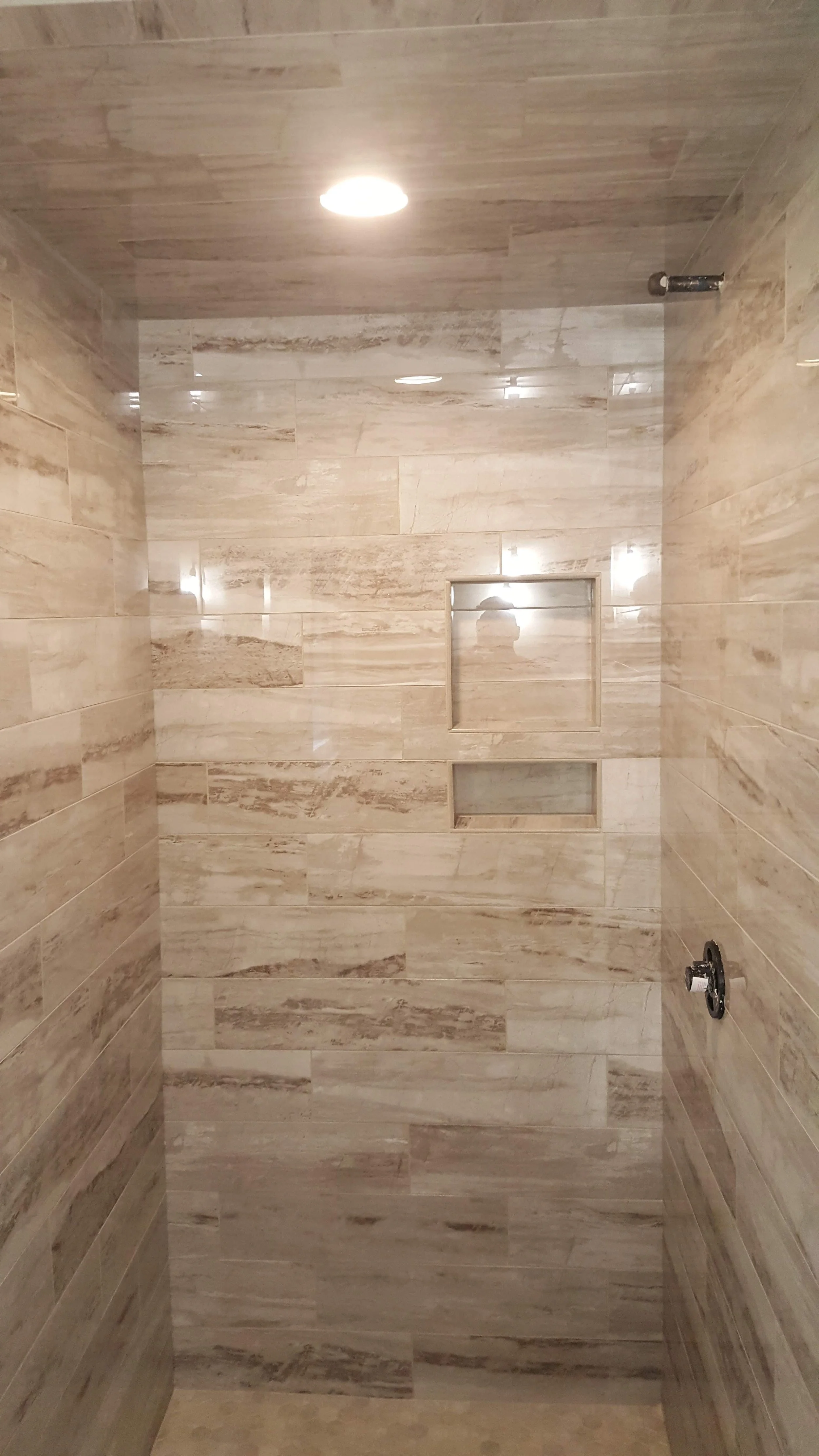 A shower with beige and light brown tiled walls and ceiling, central light, two built-in niches, and shower fixtures with no showerhead installed.