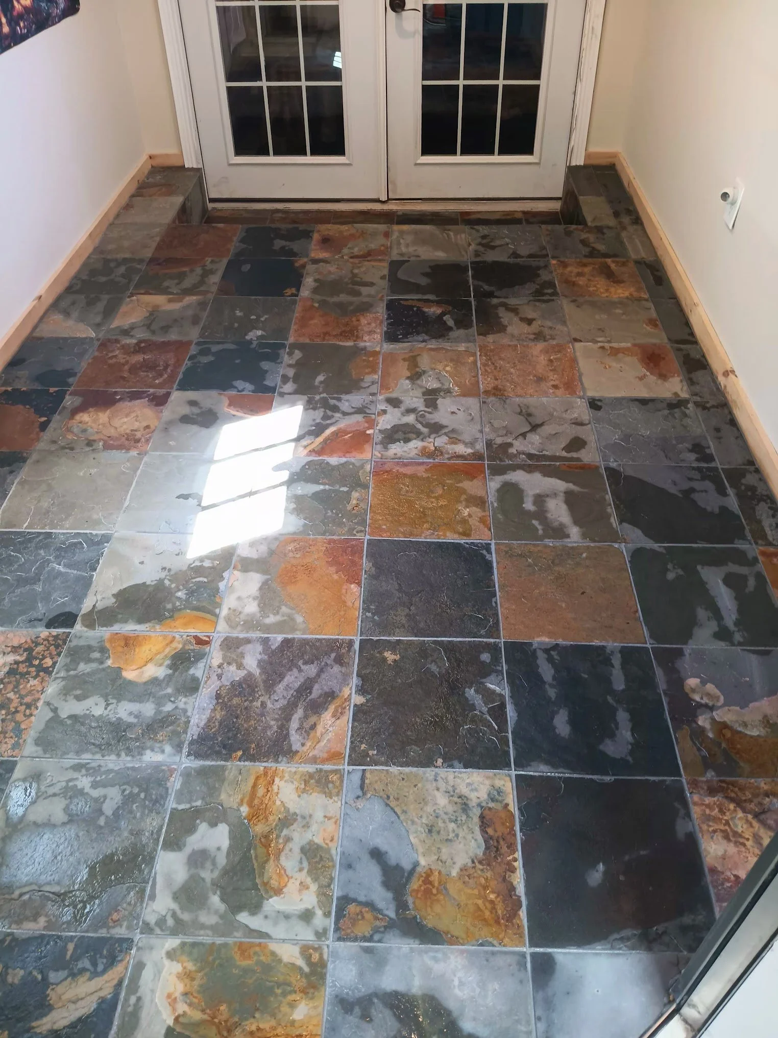 A tiled floor near a glass double door with wooden trim, featuring multicolored natural stone tiles in shades of gray, black, brown, and tan.