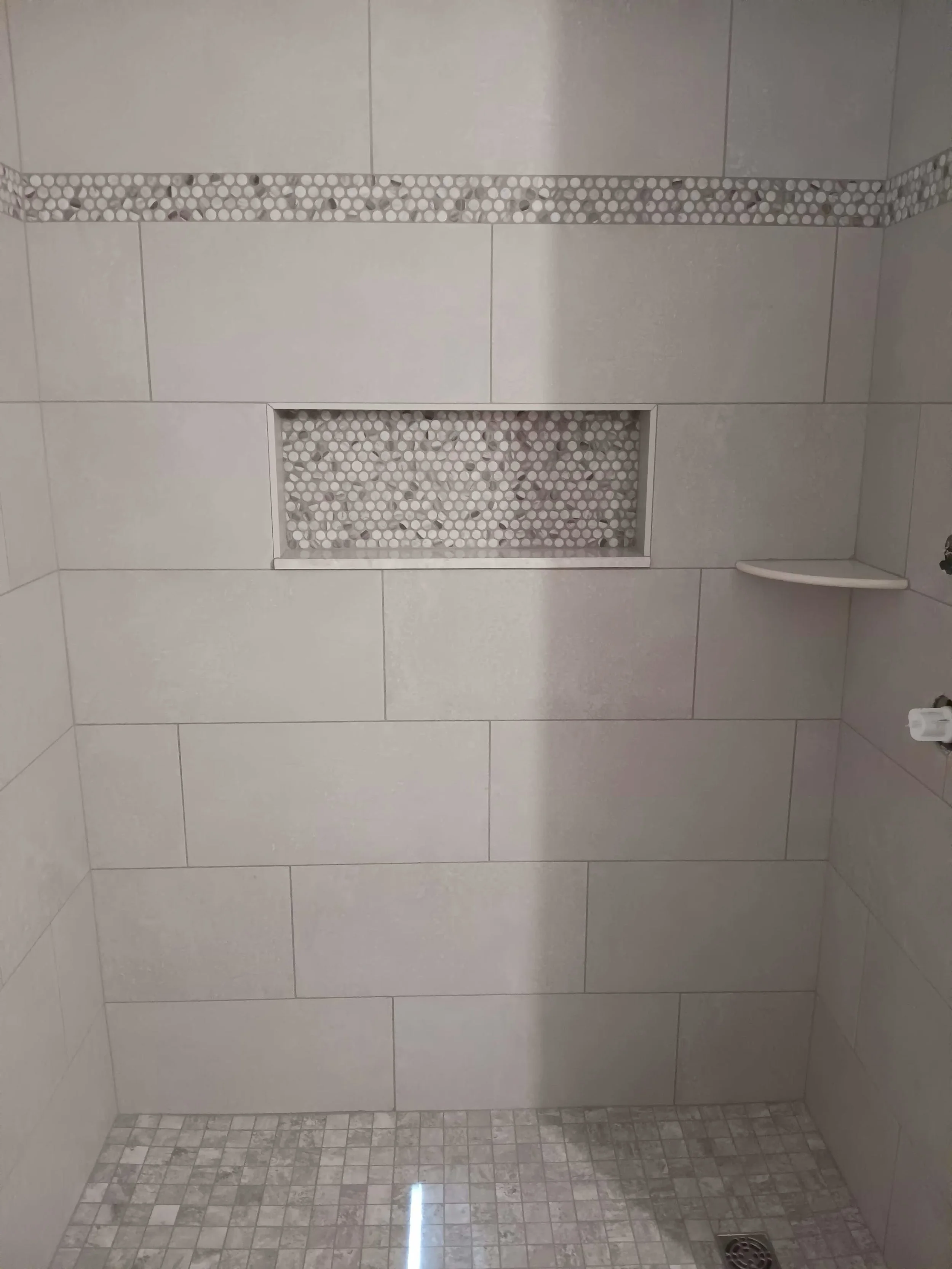 Close-up of a tiled shower wall with decorative pebble tile accents and a built-in niche holding smooth white pebbles. The bottom floor has small mosaic tiles and a drain.