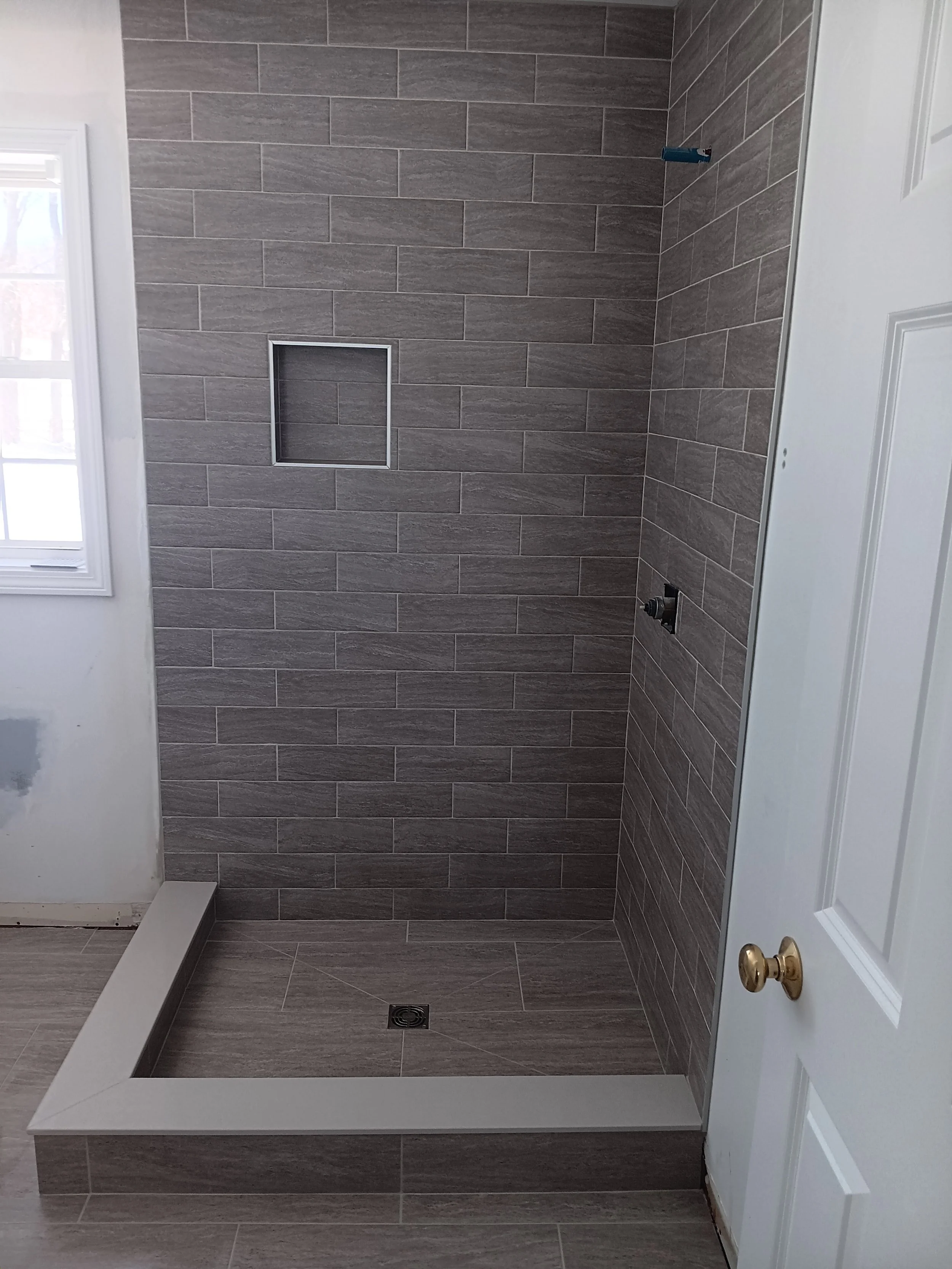 A tiled bathroom shower with brownish-gray rectangular tiles, a built-in niche, a drain on the floor, and a white door with a golden doorknob partially visible to the right.