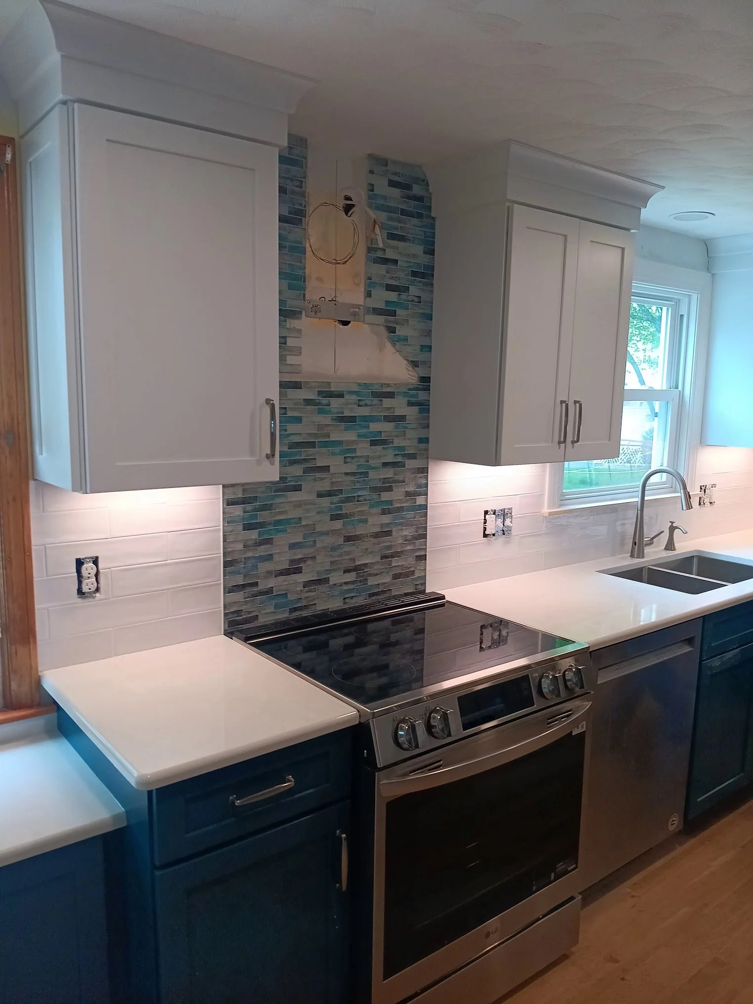 Kitchen with white cabinets, a backsplash with blue and gray tiles, stainless steel stove, black oven, white countertop, and a window above the sink.