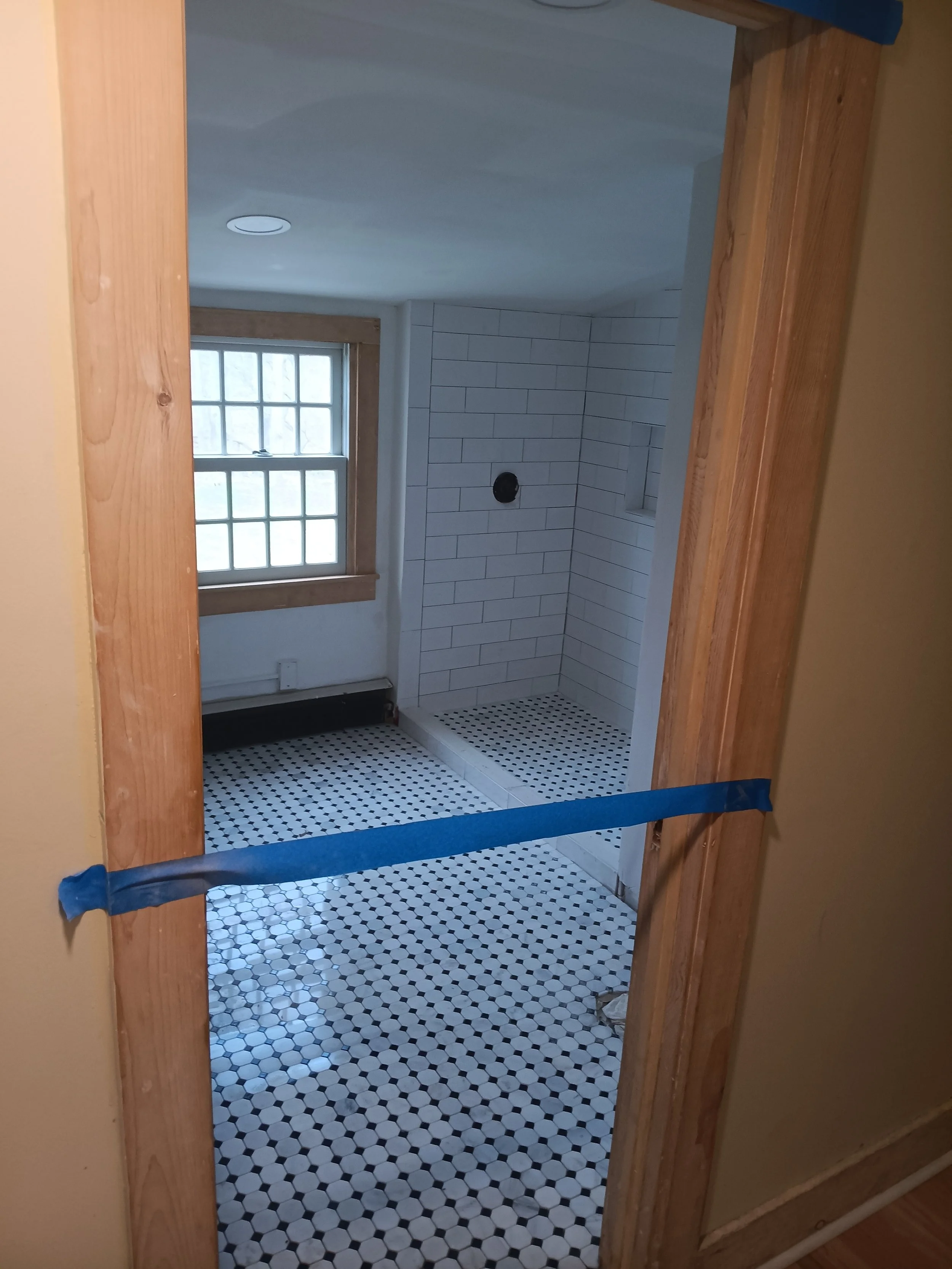 Bathroom with black and white hexagonal tiled floor, white tile shower with niche, window with wooden trim, and wall paint with a tan color.
