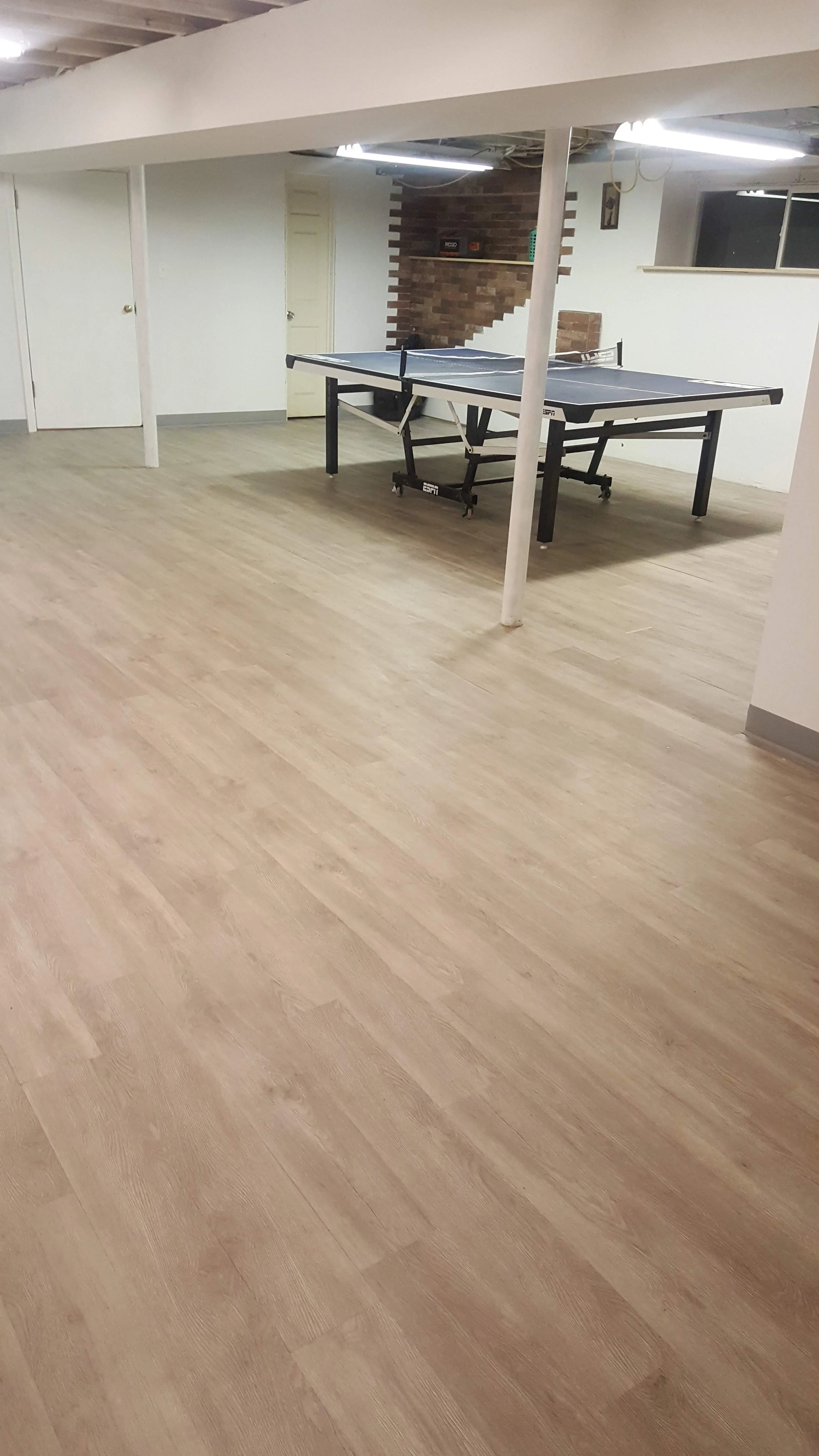 An indoor room with light-colored wooden flooring, a ping pong table in the center, white walls, and a brick accent wall in the background. There are doors and a window visible.