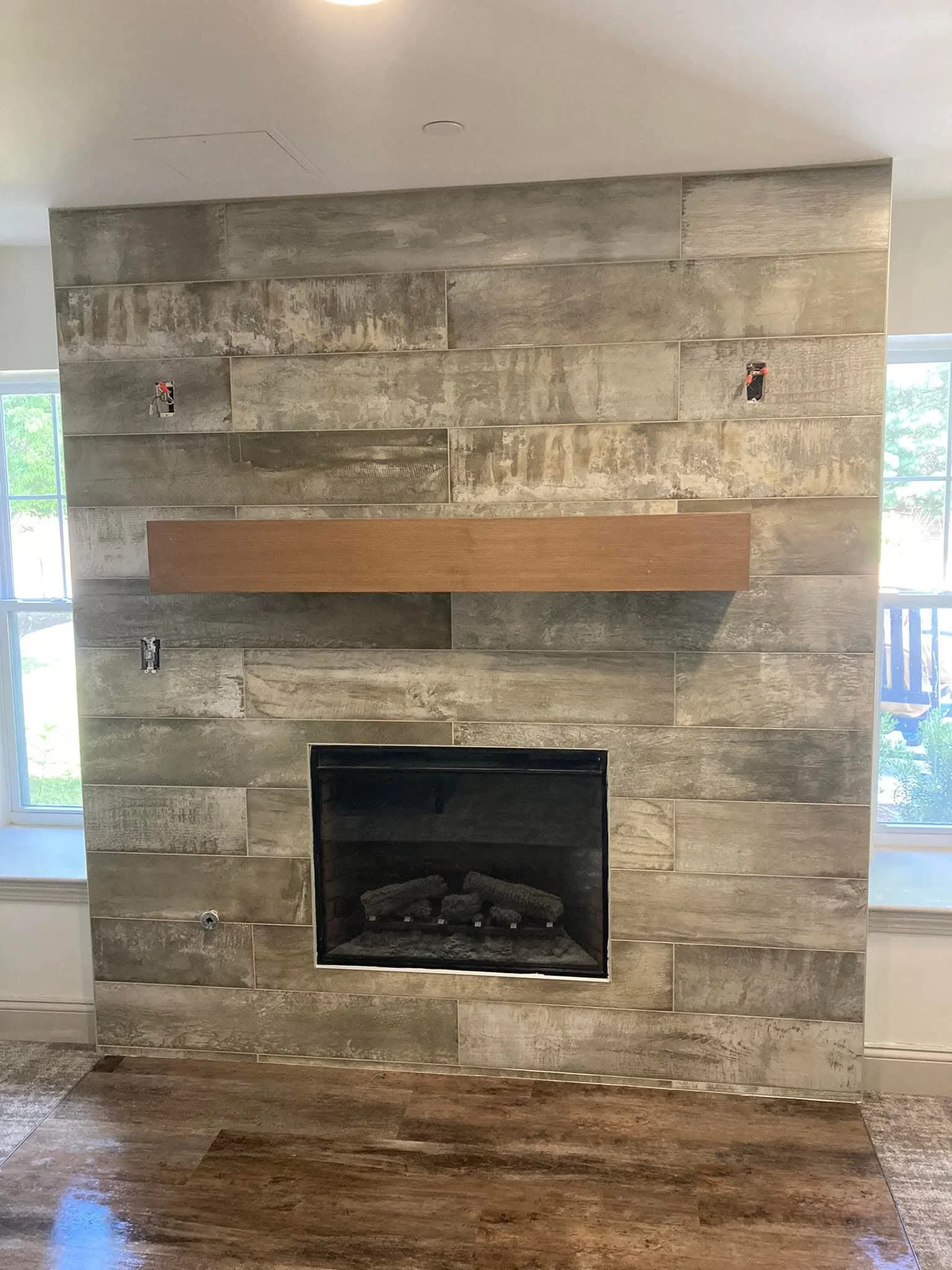 Living room with a new tiled fireplace, a wooden mantel, and two large windows with a view of greenery outside.