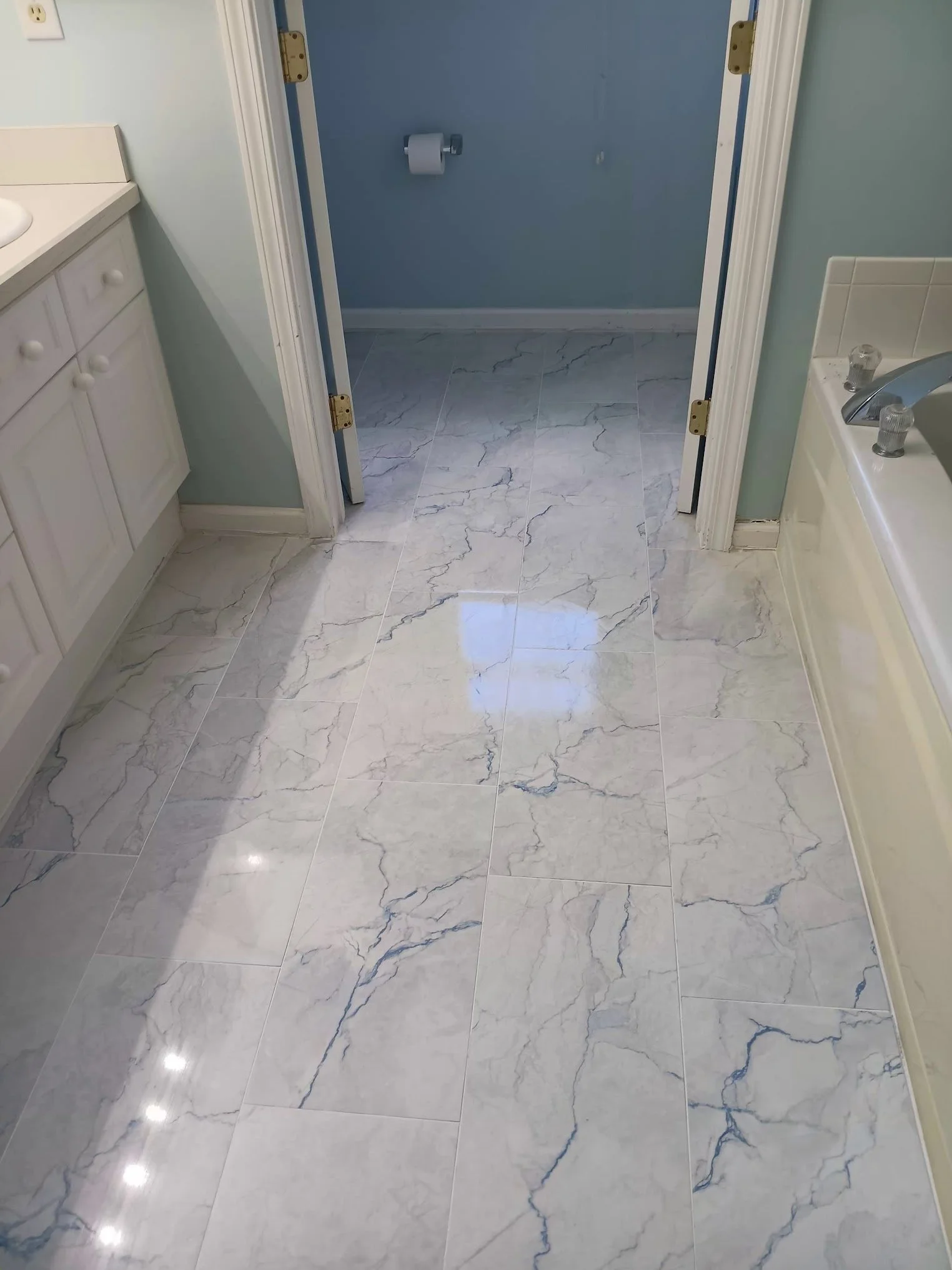 Bathroom with marble tile flooring, a closed blue door with a toilet paper holder, a white vanity with knobs on the left, a white bathtub with taps on the right, and light-colored walls.