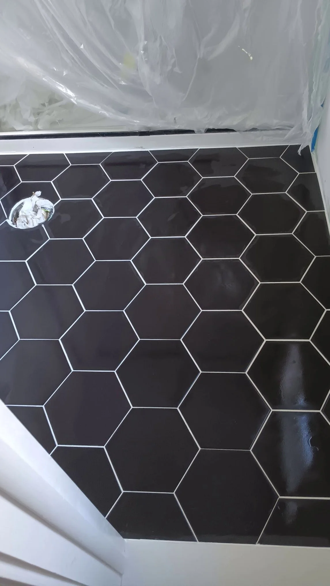 Close-up of a bathroom floor with black honeycomb tiles outlined in white, next to a shower with a plastic curtain and a drain covered with a white plastic bag.