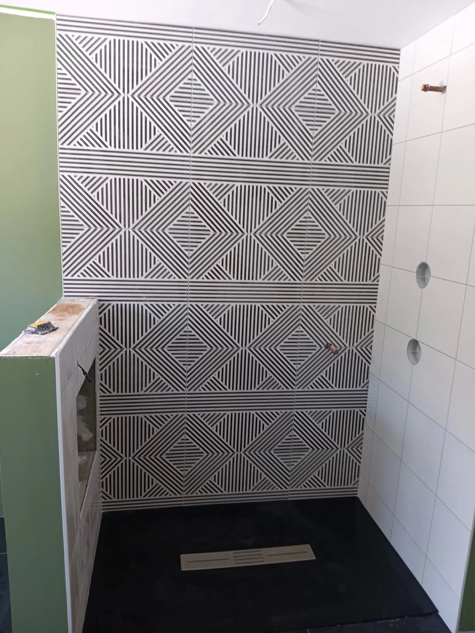 Bathroom with black and white geometric patterned tiles on the wall, white tiles on the side wall, green painted wall, and a black floor mat in front of a small bathroom vanity or sink area. There are unfinished elements and construction tools visibl