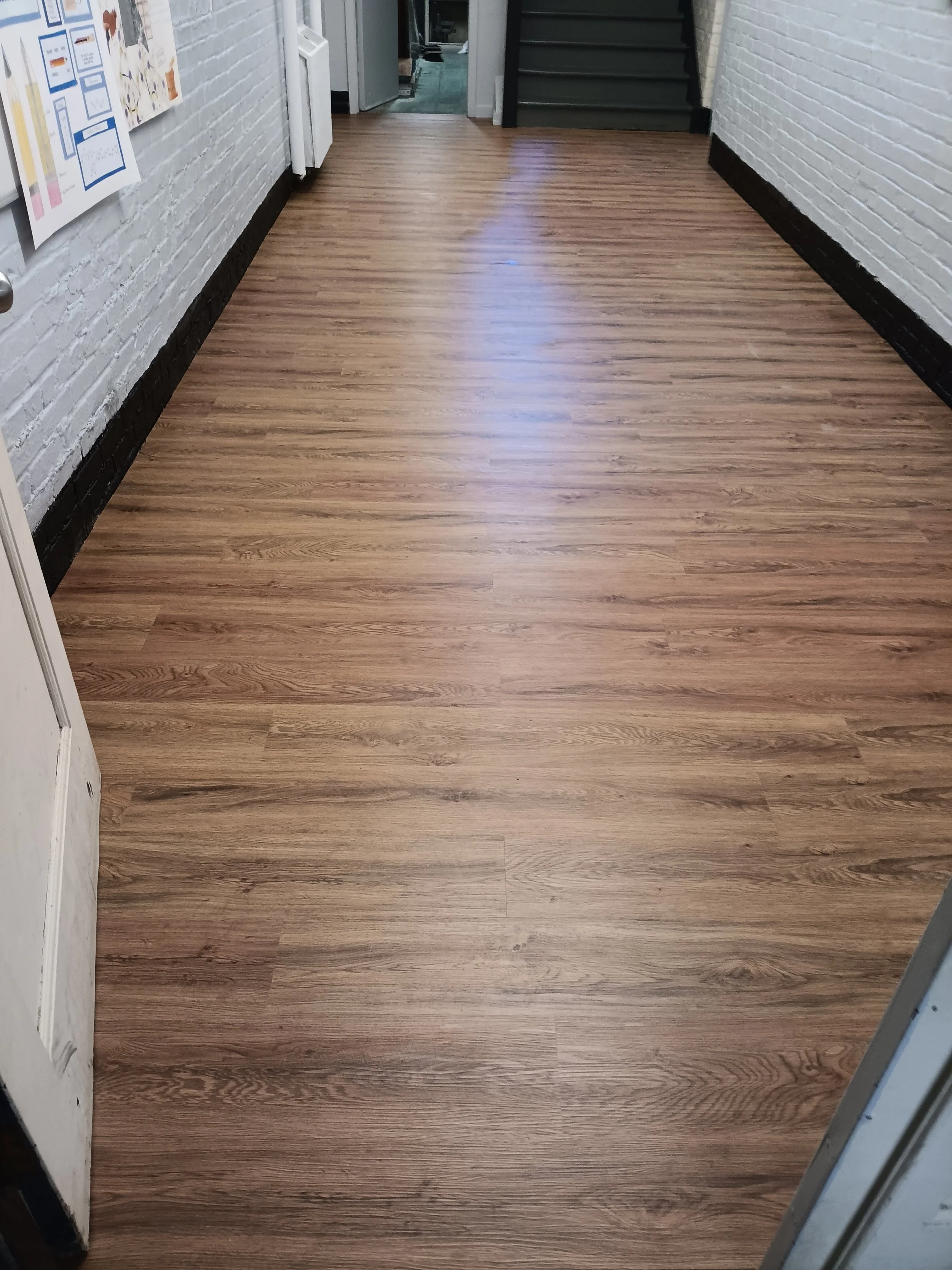 A hallway with wood flooring, white brick walls, and a staircase with dark gray steps leading up. There is a door at the end of the hallway and some posters on the wall to the left.