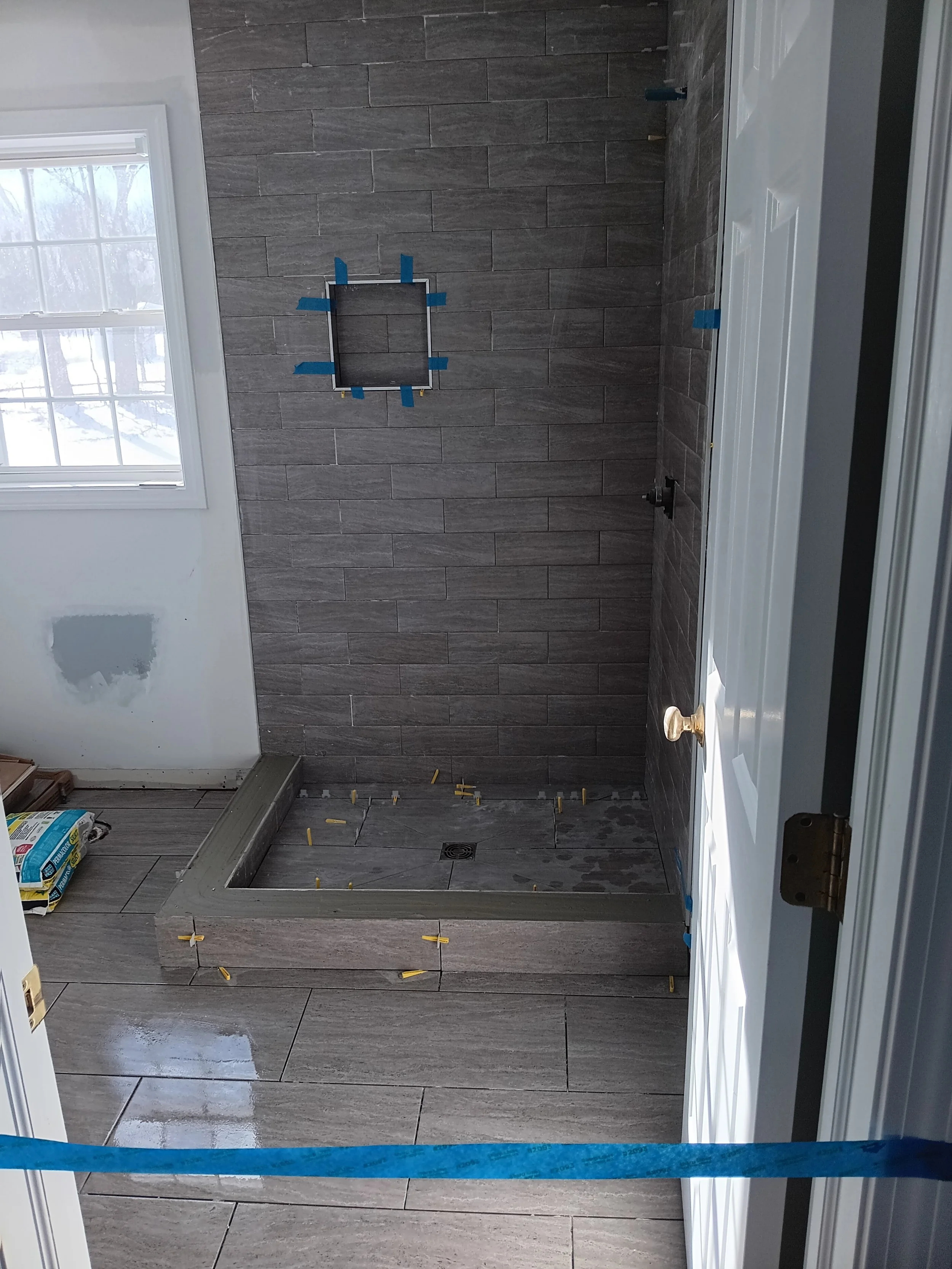 Interior of a bathroom under renovation with gray tile flooring and walls, a window on the left, a partially installed shower base, and construction tools and materials scattered around.