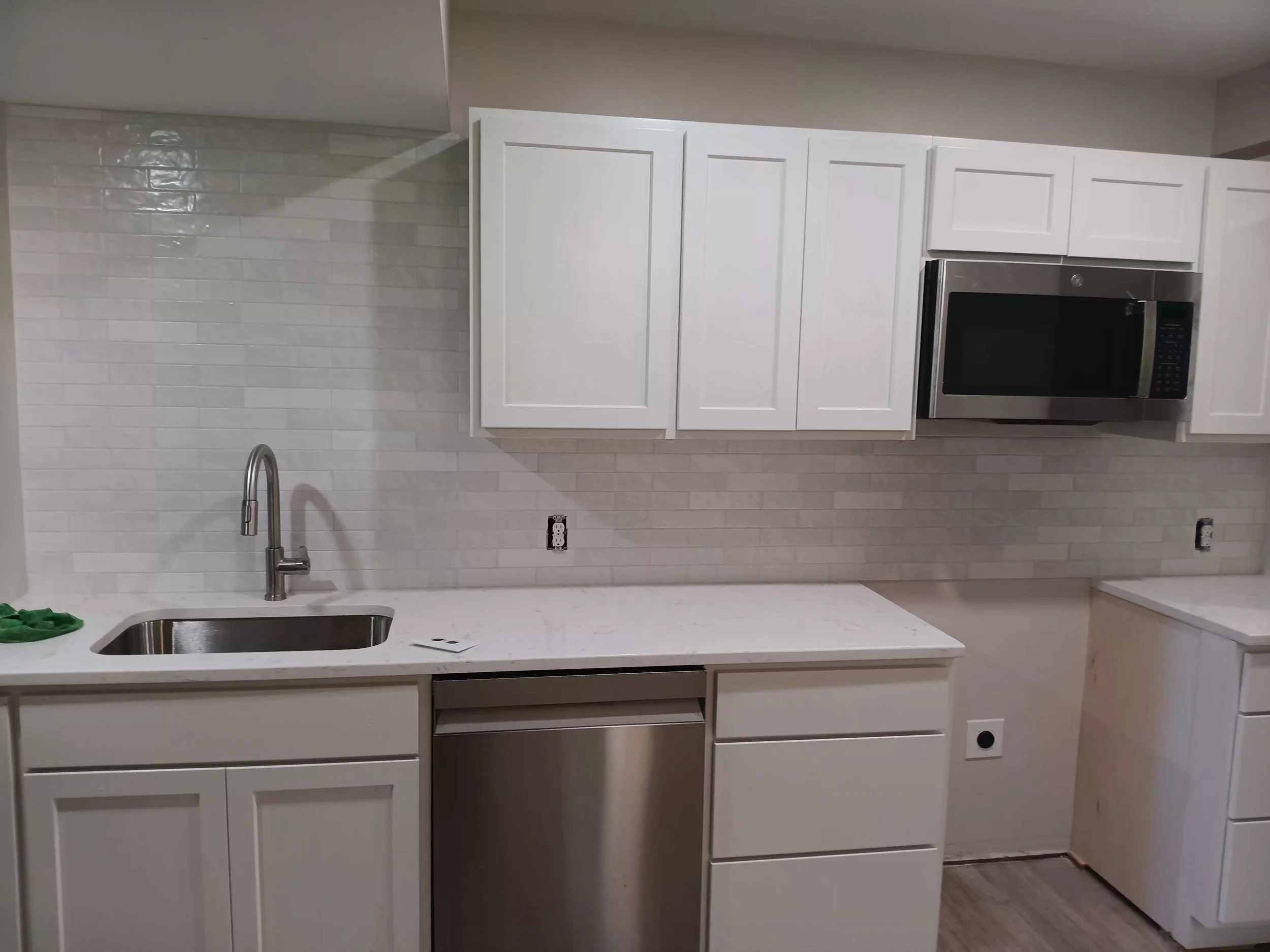 A modern kitchen with white cabinets, a stainless steel microwave, a white countertop with a sink and faucet, and a white brick-style backsplash.