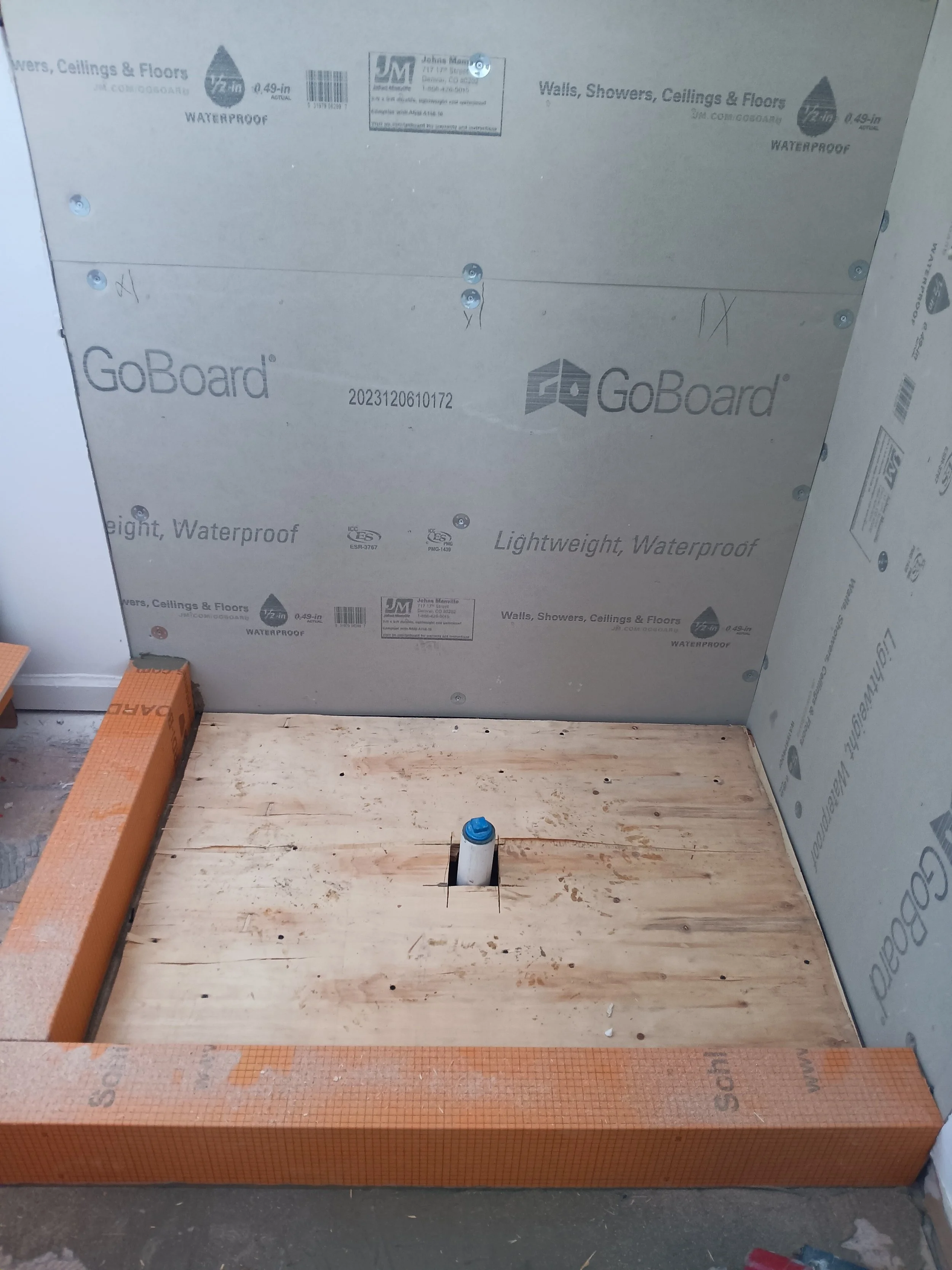 Construction area with a wooden platform, a pipe protruding from the floor, and waterproof wall panels installed on three sides.