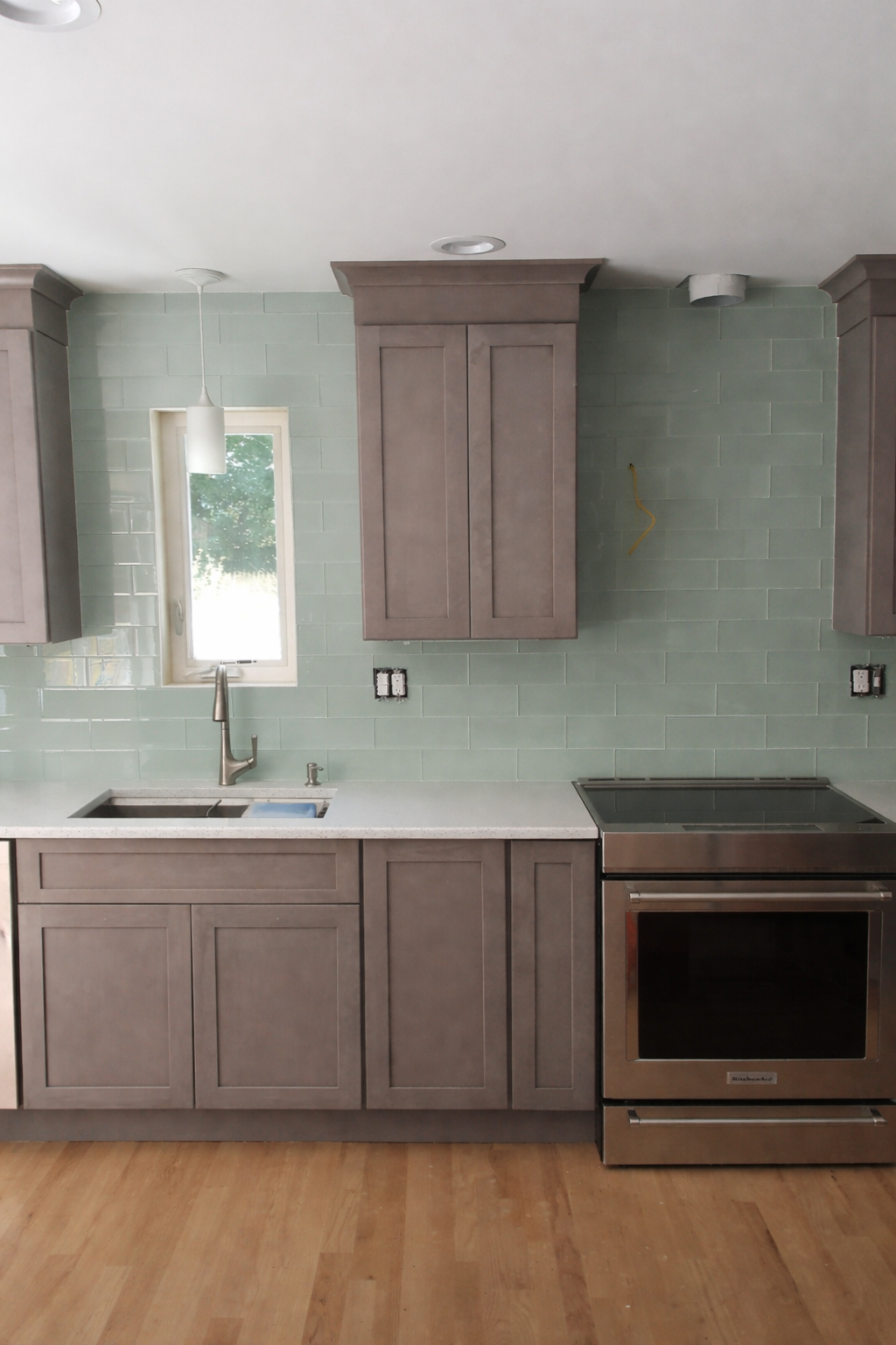 A modern kitchen with gray cabinets, a white countertop, a stainless steel oven, a single window, and green tile backsplash.
