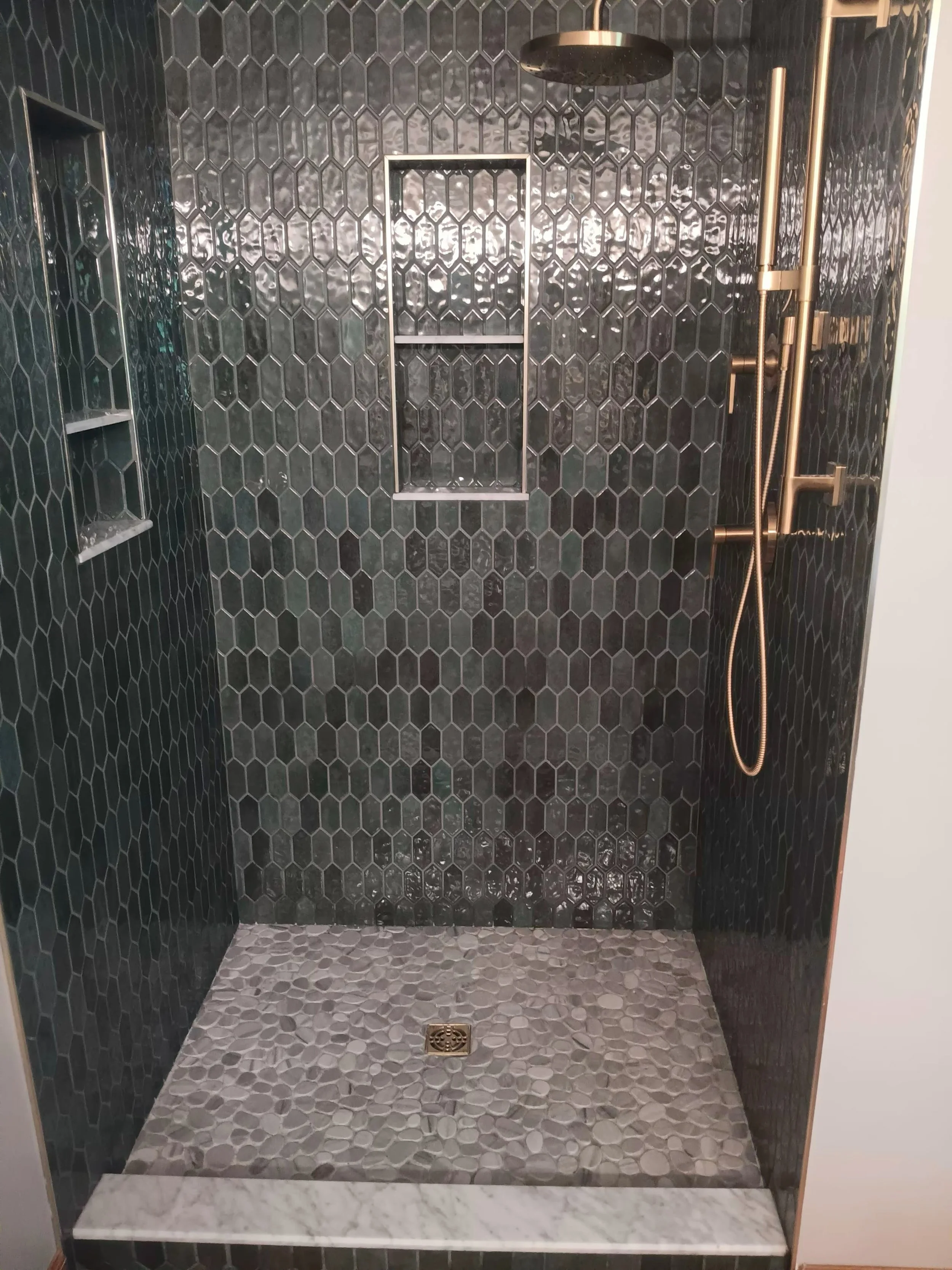 A modern walk-in shower with black hexagonal tiles on the walls and pebble-patterned tiles on the floor. It features a rainfall showerhead, a handheld shower, a built-in niche, and a marble threshold at the entrance.