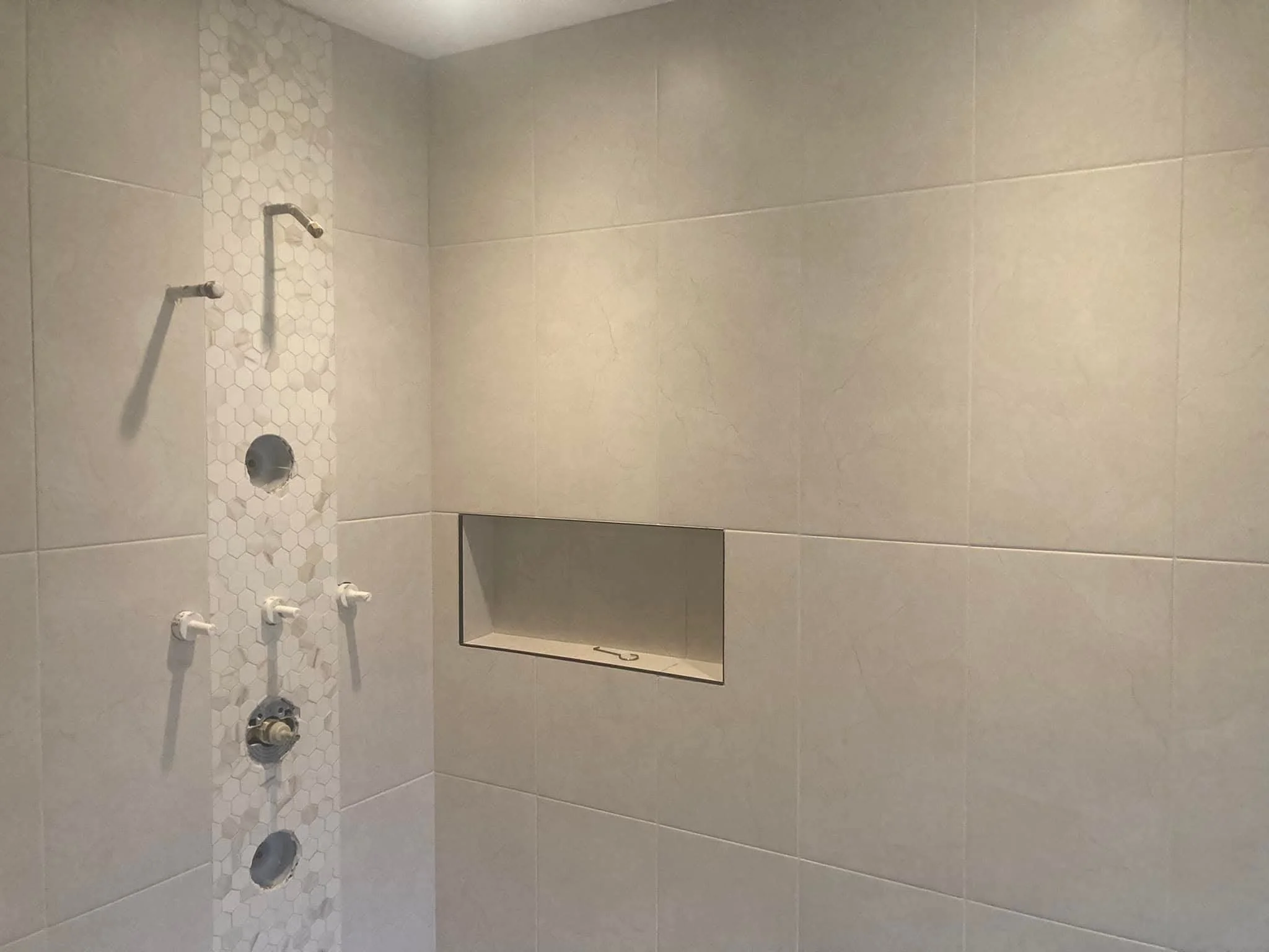 Shower area with beige tiles, a vertical strip of small hexagonal tiles, multiple showerheads and controls, and a built-in niche for toiletries.