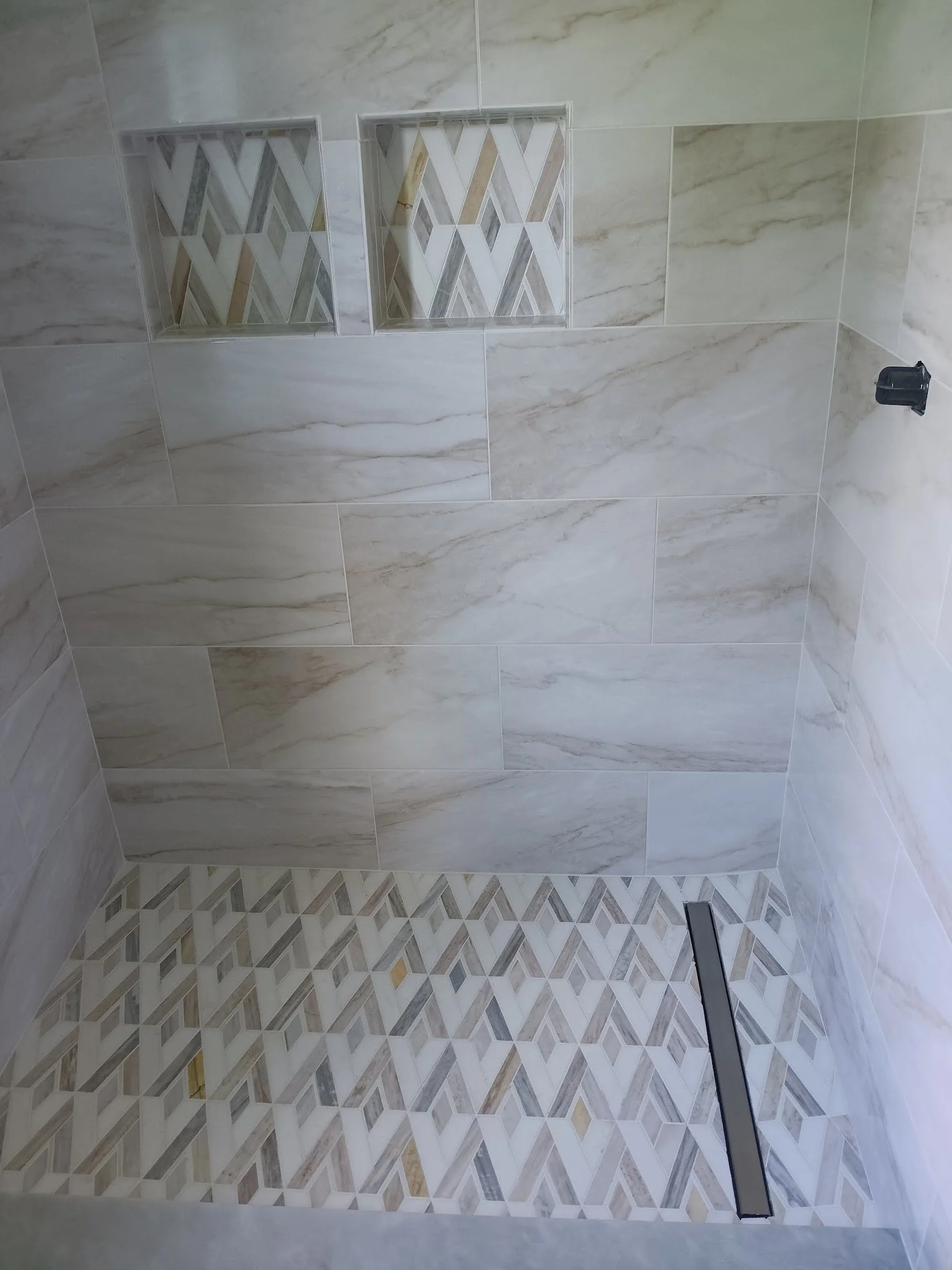 A bathroom shower with a marble tile wall and decorative accent tiles in a geometric pattern on the shower floor and interior wall. There is a black shower valve handle on the right side.