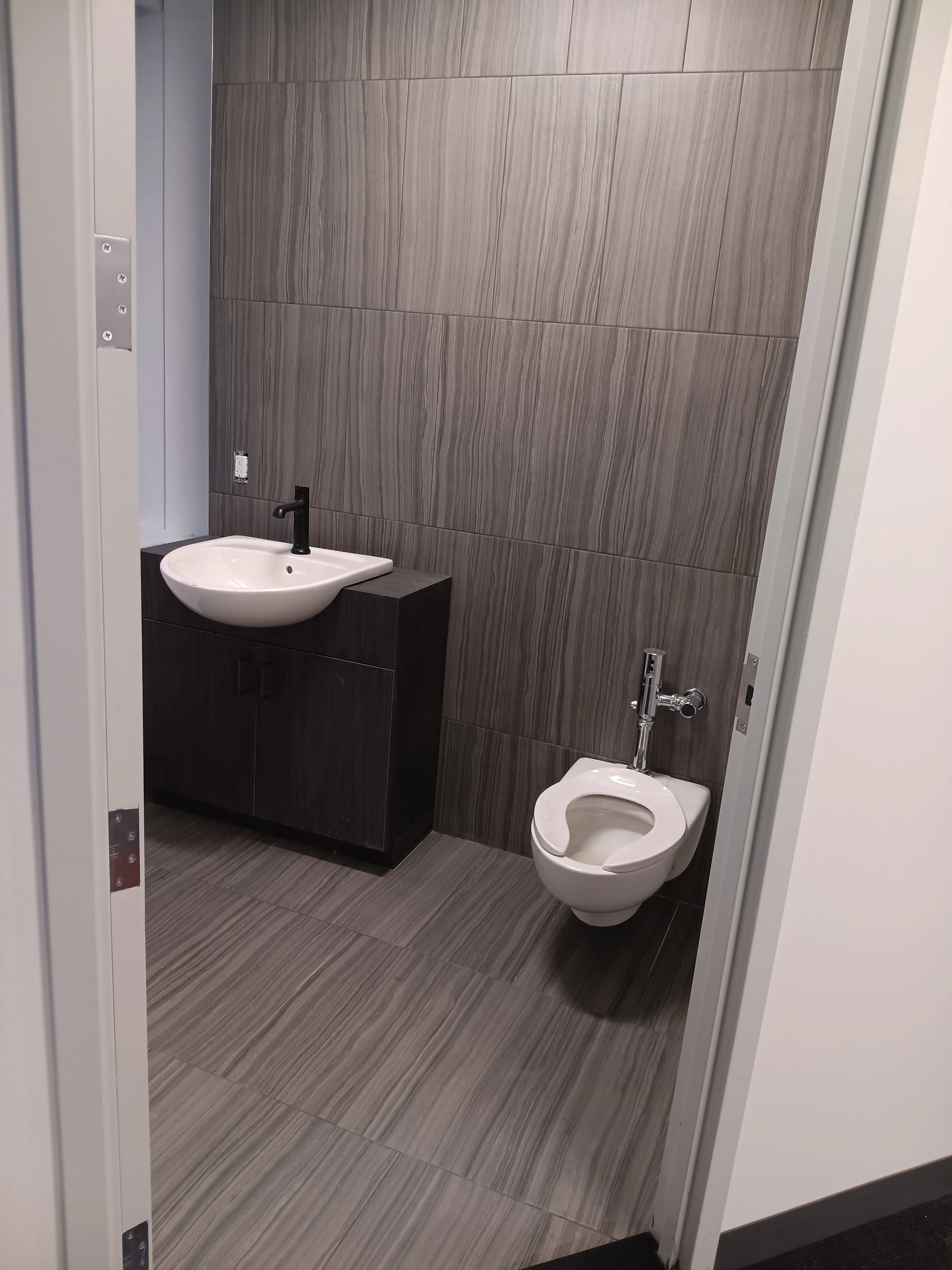 A small restroom with gray tiled walls and floor, a white sink with a black faucet on a dark wood cabinet, and a white toilet with a modern flush tank.