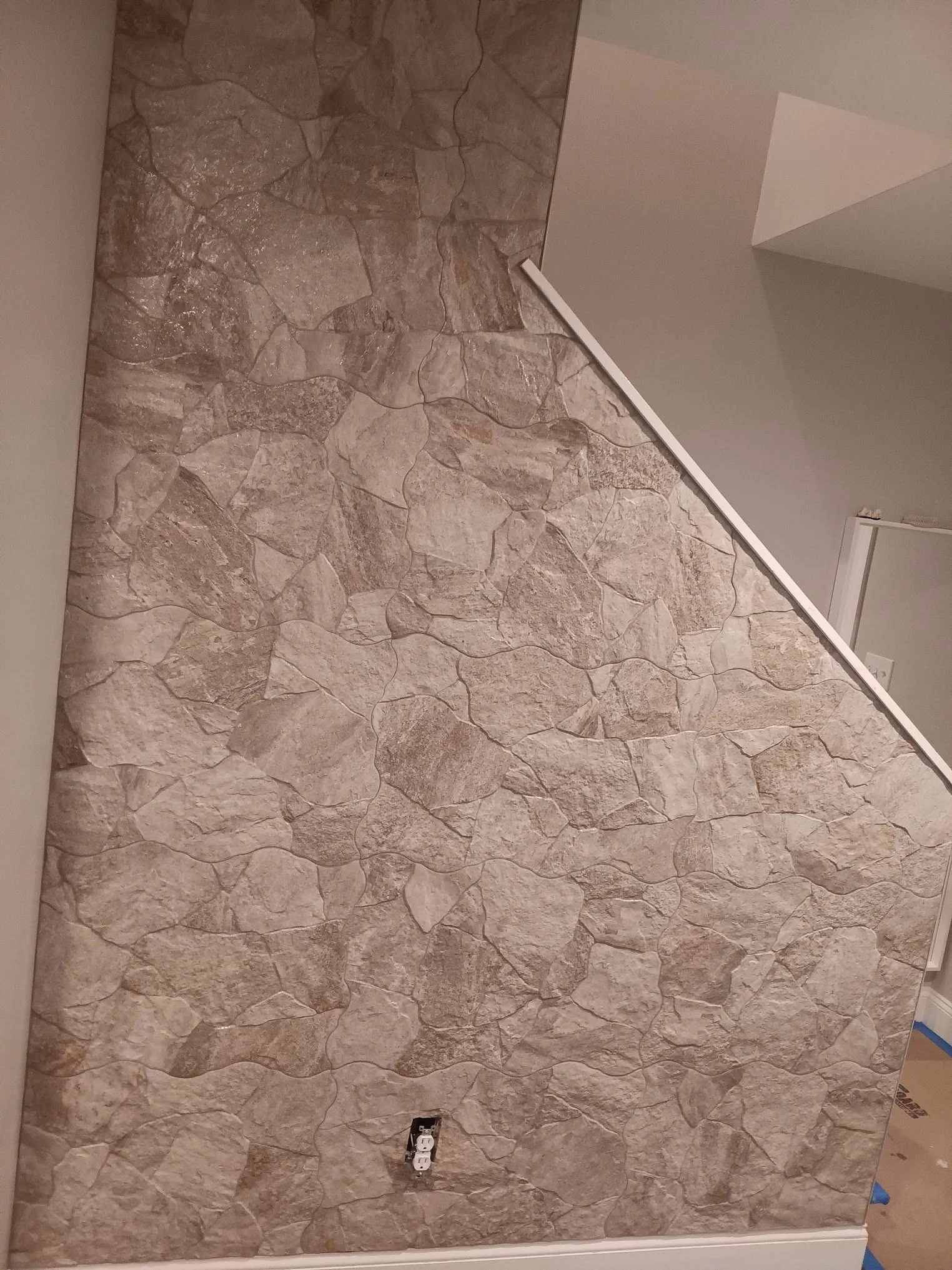 Partially finished wall with stone-like textured wallpaper, electrical outlet, and white trim at the bottom.
