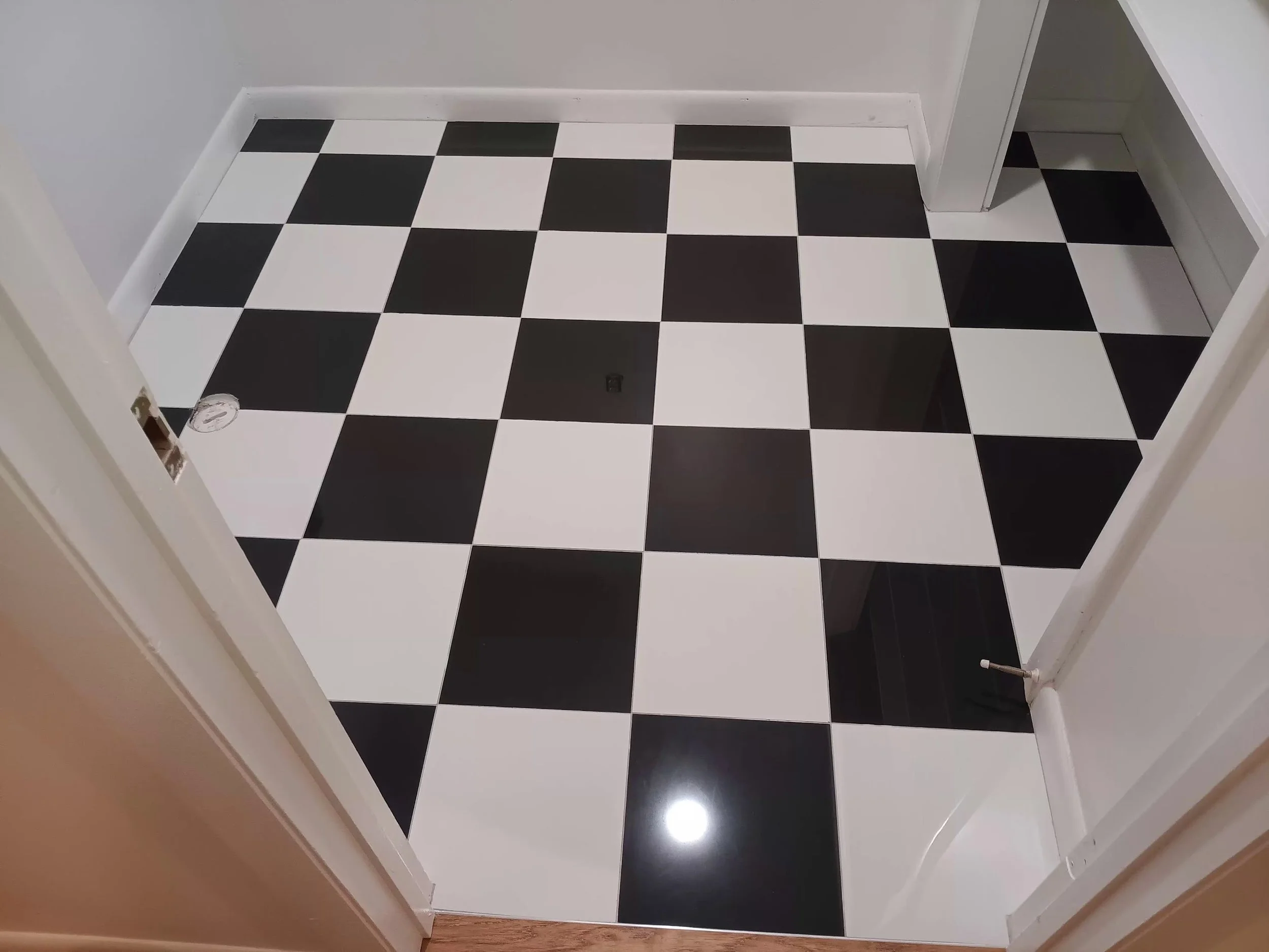 A black and white checkered tile floor in a room with white walls and baseboards, and a doorway leading to another room.