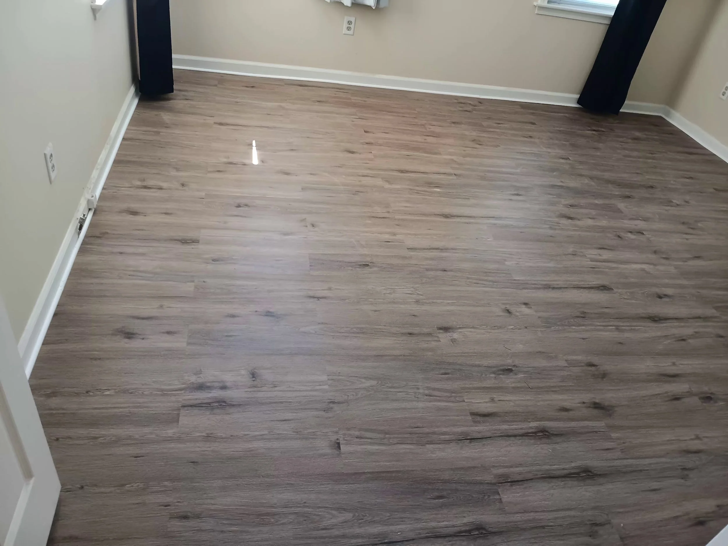 A room with wood laminate flooring, beige walls, white baseboards, a white electrical outlet, and a window with black curtains.