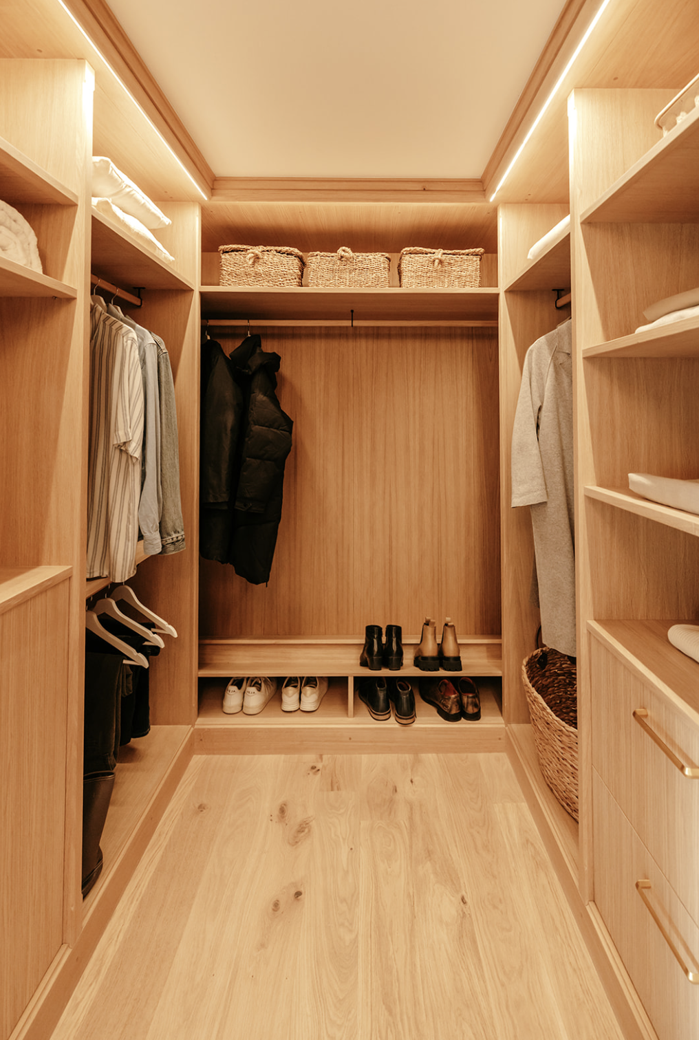 Un dressing en chêne clair avec des étagères contenant des vêtements pliés, des chaussures, des sacs et des baskets, avec un éclairage intégré au plafond. Dressing réalisé par l'Atelier Set à Perros-Guirec.