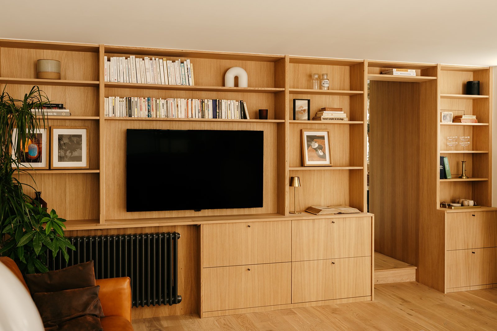 Une bibliothèque en chêne clair avec des étagères remplies de livres, une télévision noire au centre, des cadres avec des photos, des objets décoratifs et une plante verte à gauche. Bibliothèque réalisée par l'Atelier Set à Perros-Guirec.