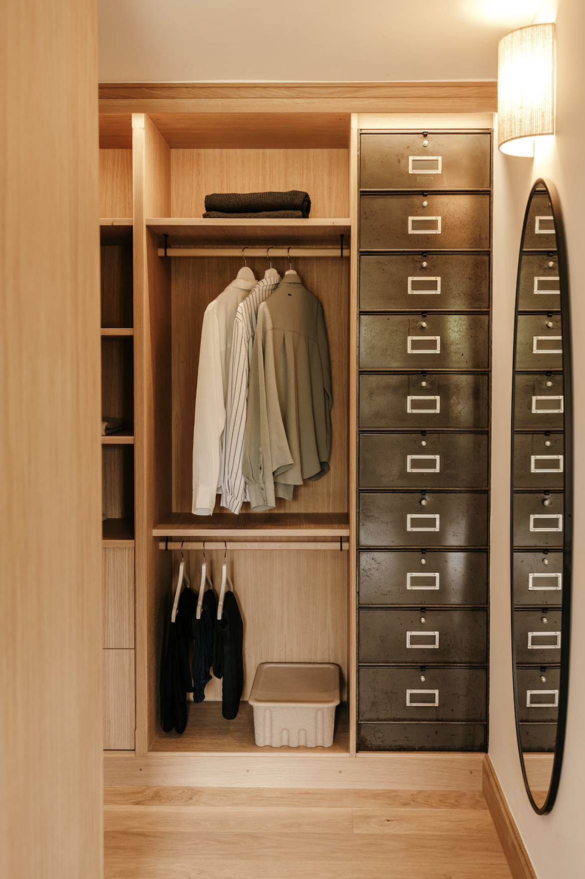Dressing en bois avec des vêtements suspendus, une pile de vêtements, une petit meuble d'archives avec des tiroirs en métal, un miroir et une lampe. Dressing réalisé par l'Atelier Set à Perros-Guirec.