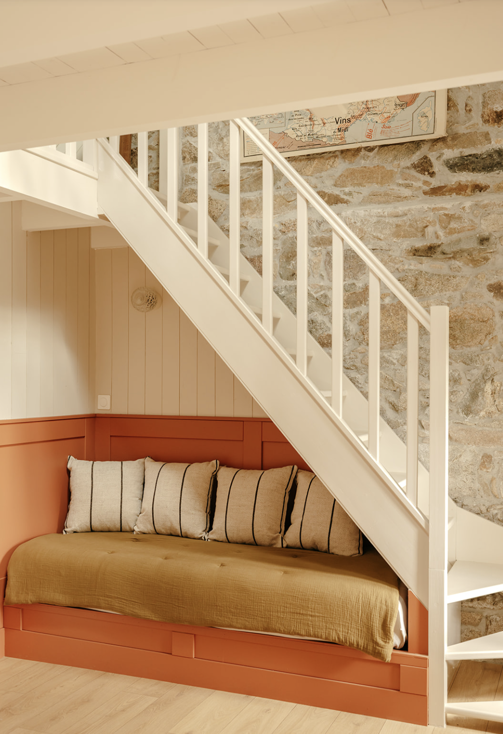 Coin salon avec banquette en bois orange, coussins beige avec rayures noires, escalier blanc en bois, mur en pierre et carte scolaire vintage accrochée au mur.