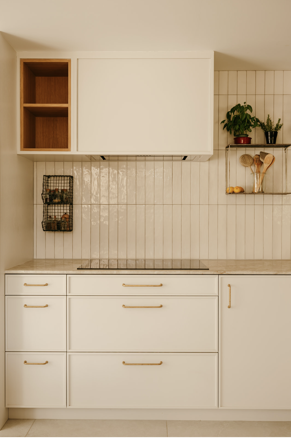 Cuisine moderne avec des tiroirs blancs, des poignées en lai, un plan de travail en pierre clair, des étagères en métal avec des plantes aromatiques et des ustensiles de cuisine, et un mur recouvert de carreaux blancs.