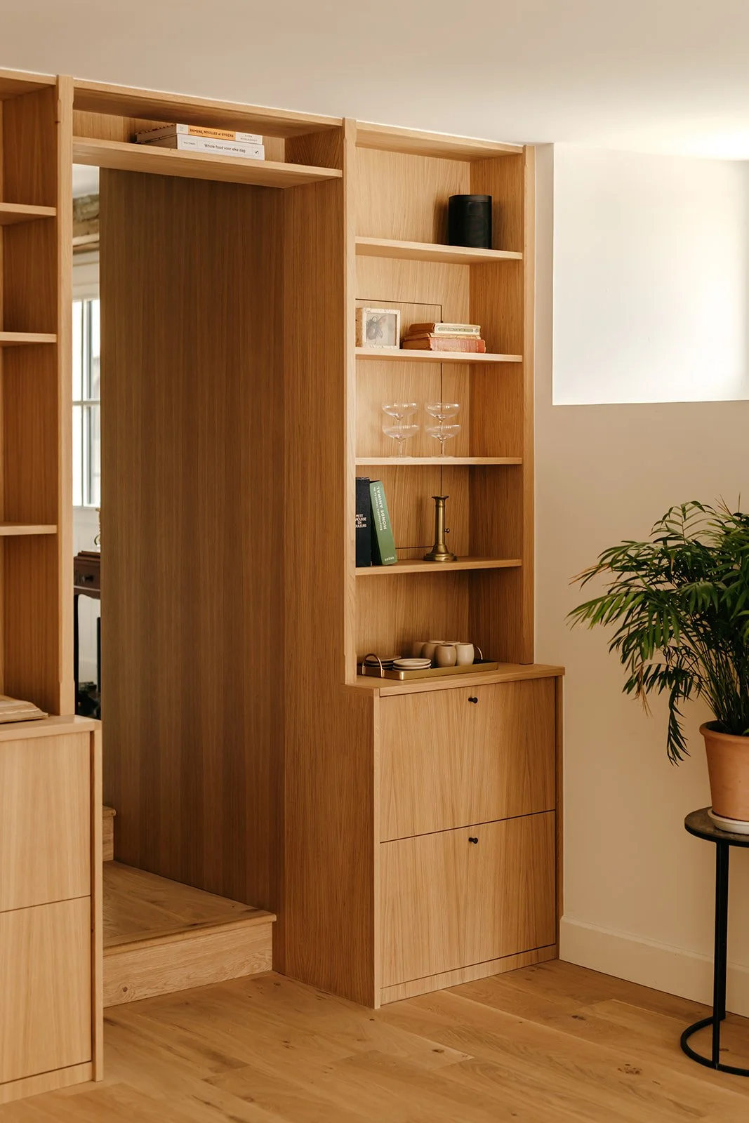 Étude d'une étagère en bois clair avec des livres, des verres à cocktail en verre, une bougie, des tasses, et une plante verte à côté.