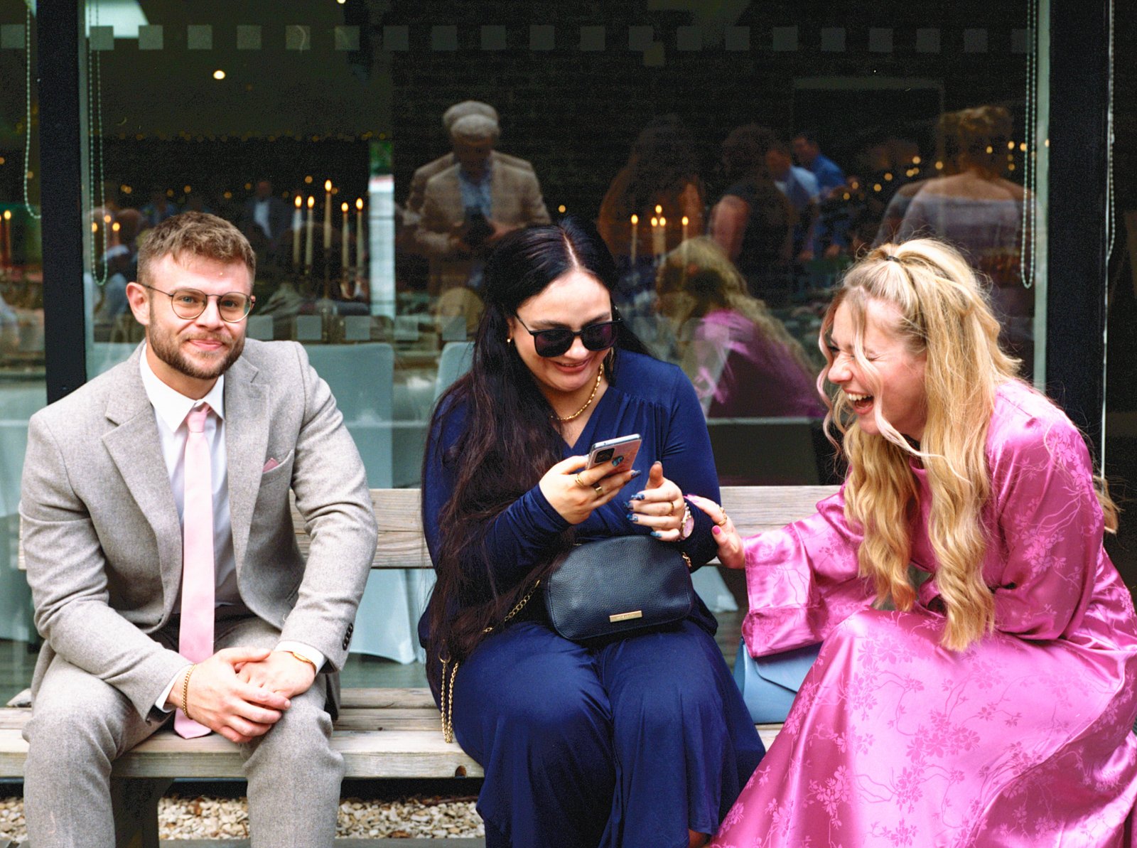 Three people sitting on a bench outside, laughing and smiling while looking at a phone.