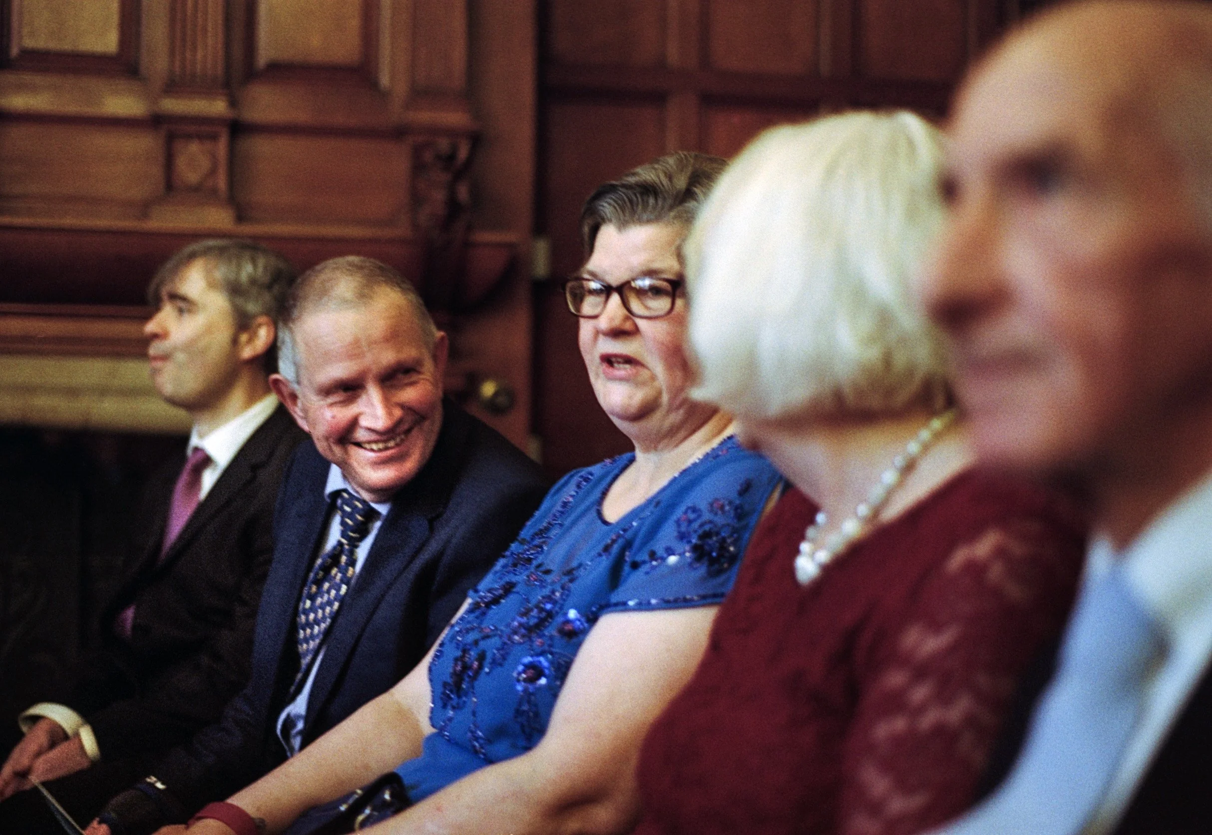 A group of well-dressed people sitting in a row at a formal event, engaged in conversation and smiling.