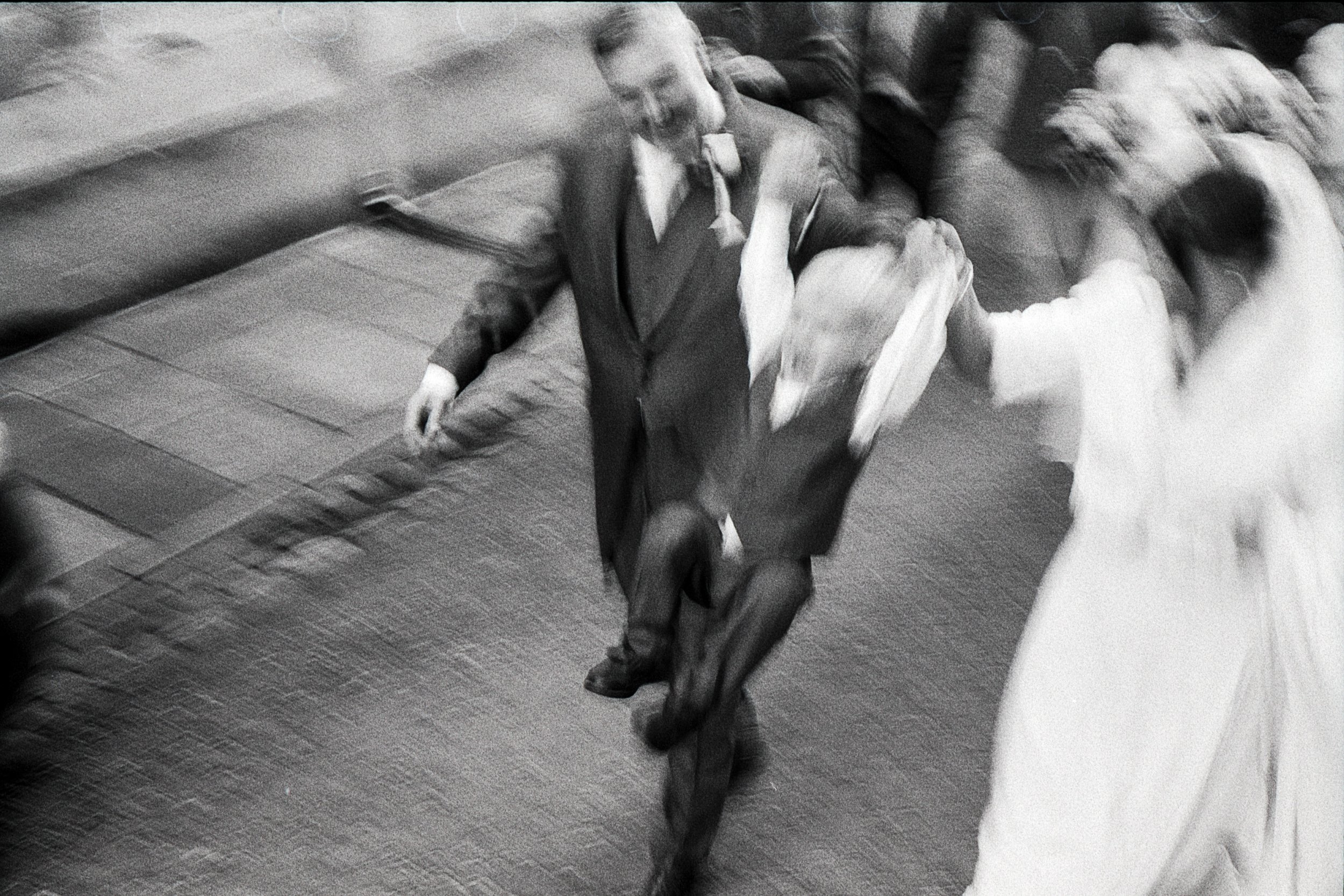Black and white photo of a man and woman dancing together, holding hands, with other people in the background.