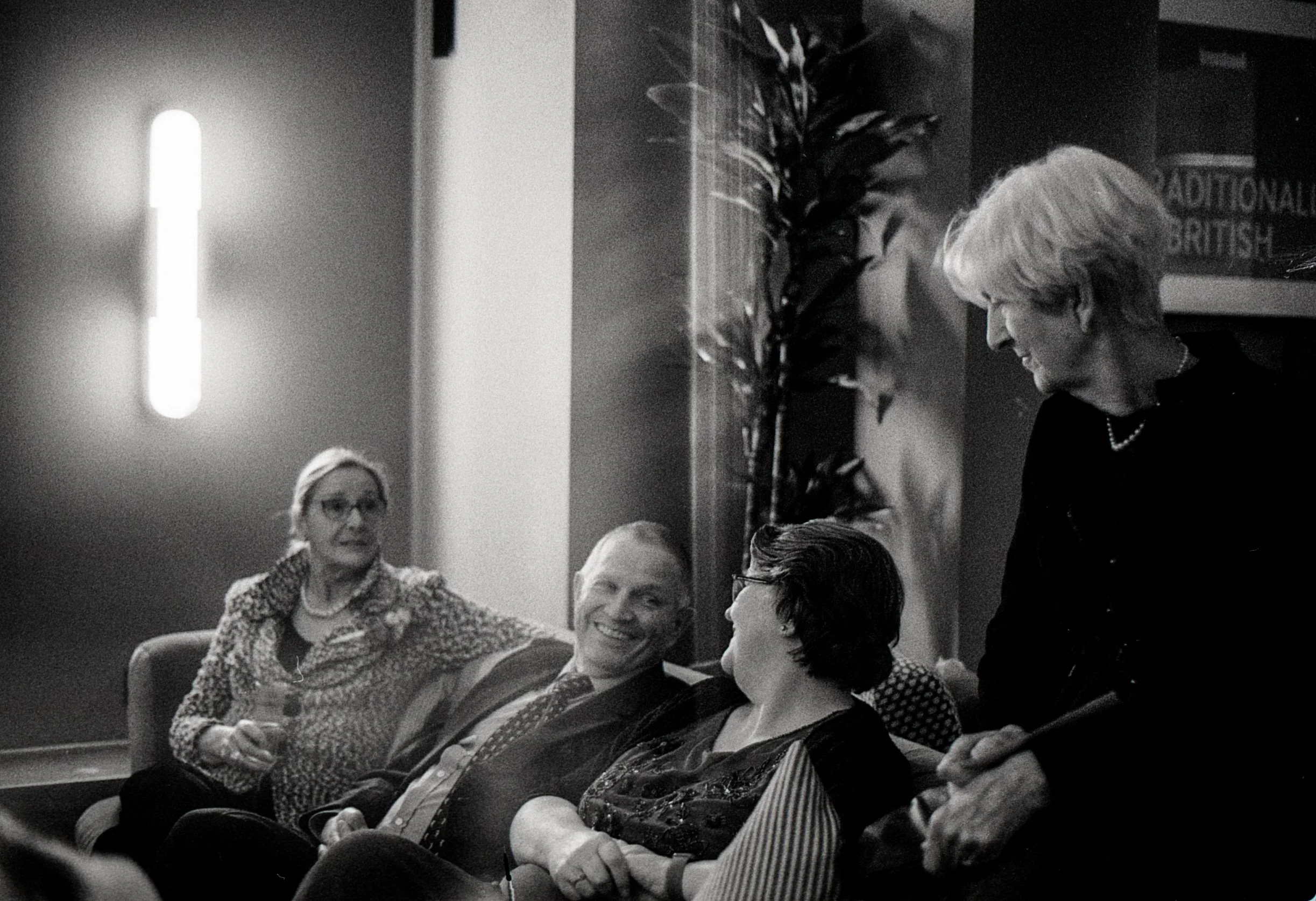 Four people, three women and one man, sitting in a room engaged in conversation, with one woman standing and smiling at the others. The woman standing has short hair and is holding a pen.