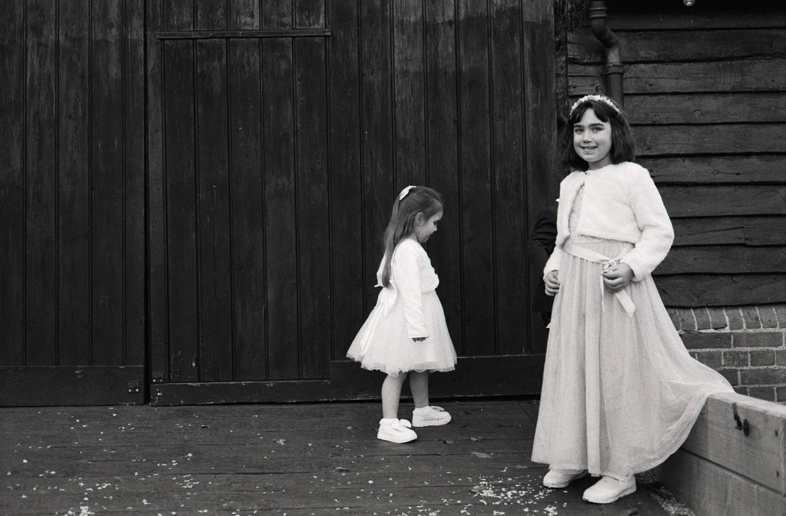 Black and white photo of two young girls dressed in fluffy sweaters and skirts, standing near a dark wooden wall and a brick wall, on a wooden floor scattered with small objects or leaves. The girl on the right smiling at the camera, the other girl l