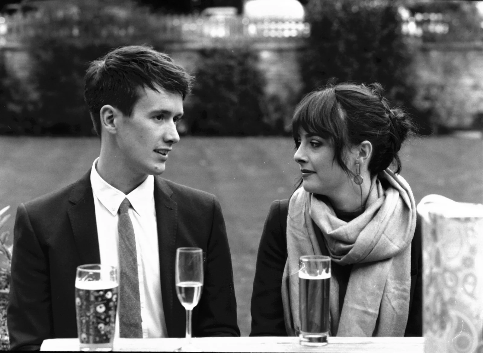 A black and white photo of a young man in a suit and a young woman with a scarf, sitting at a table outdoors with drinks, engaging in a conversation.