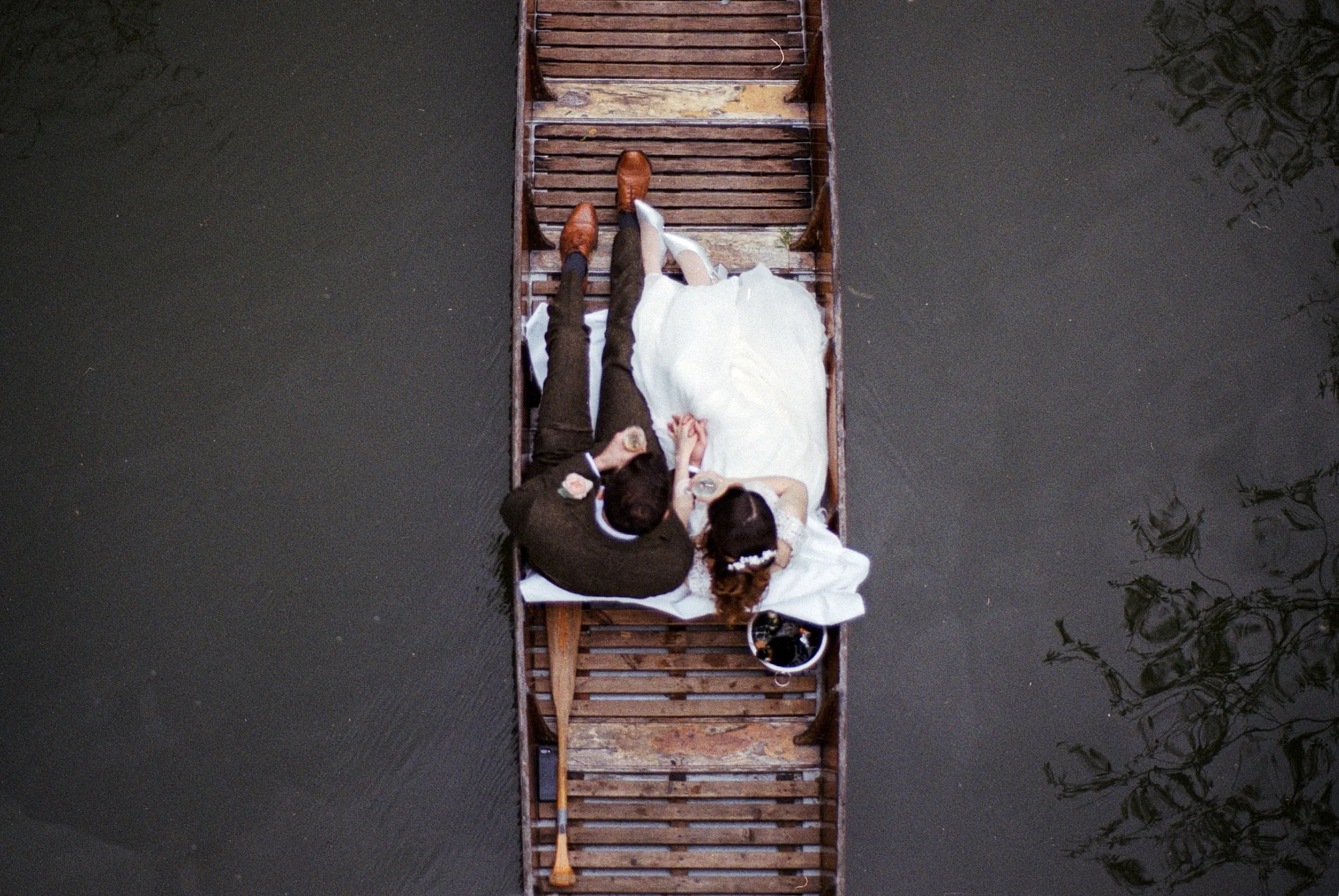 A bride and groom lying on a wooden raft on a body of water, holding hands, with the bride in a white wedding dress and the groom in a dark suit, including an oar resting on the raft.