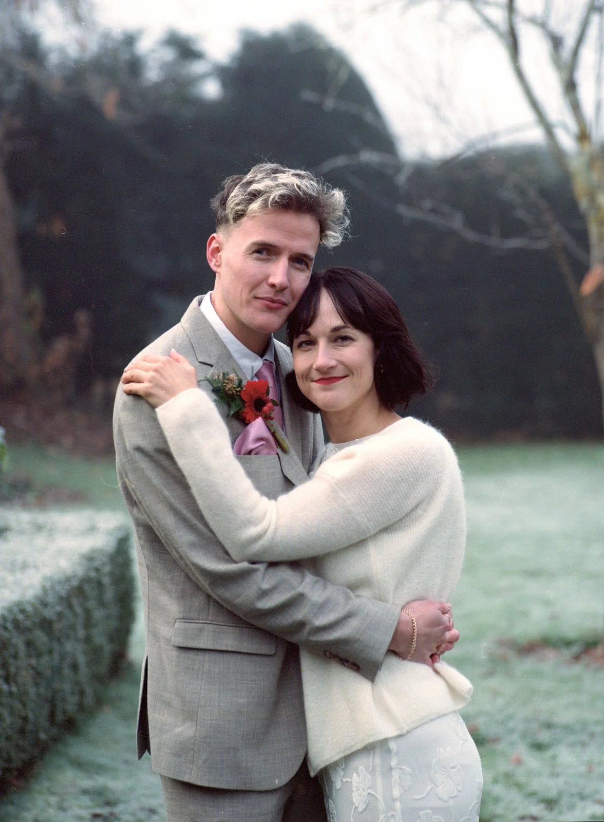 A happy couple hugging outdoors in a natural setting with trees in the background, dressed in formal attire.