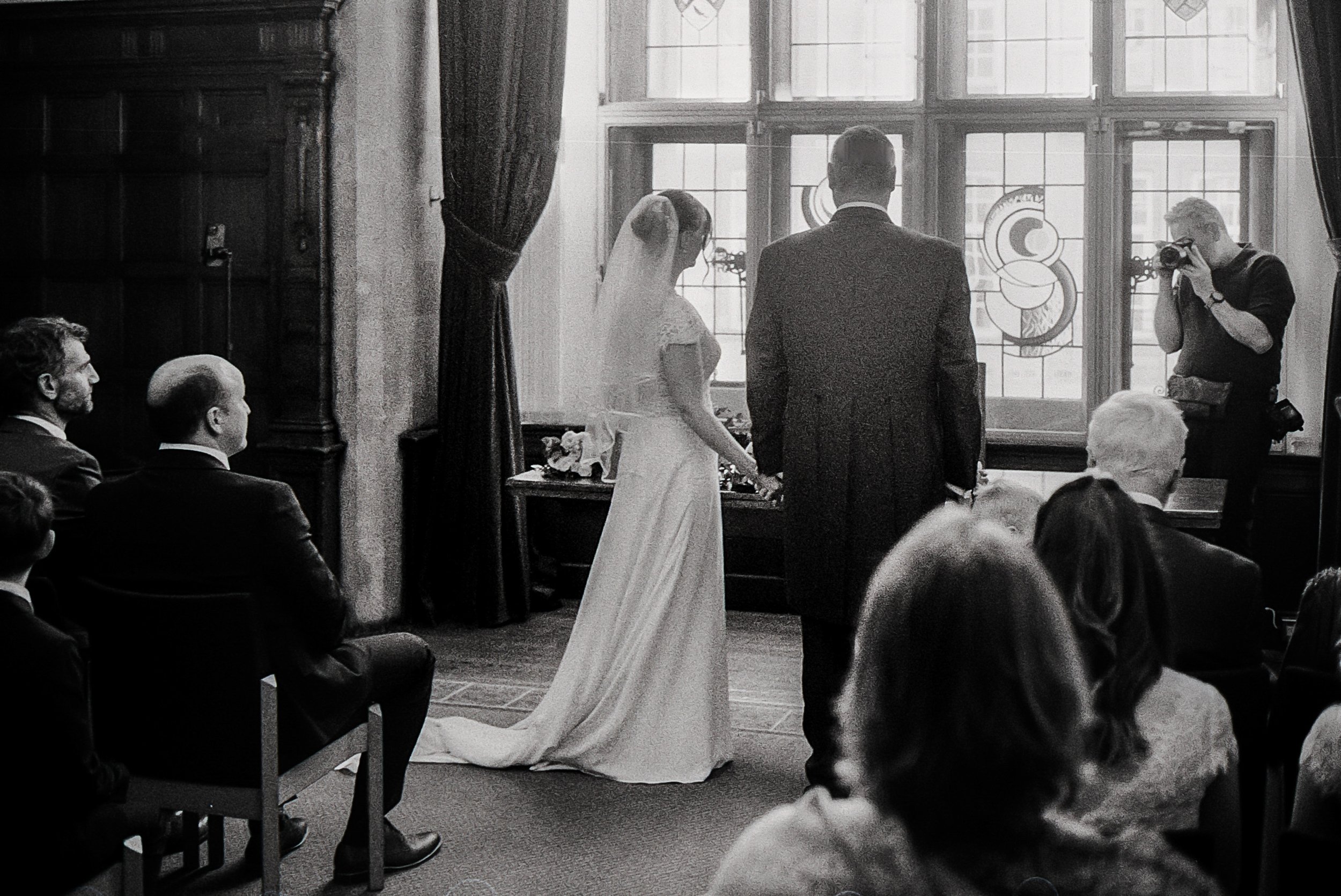 A wedding ceremony with a bride and groom holding hands in a room with large stained-glass windows, surrounded by seated guests, and a photographer taking pictures.