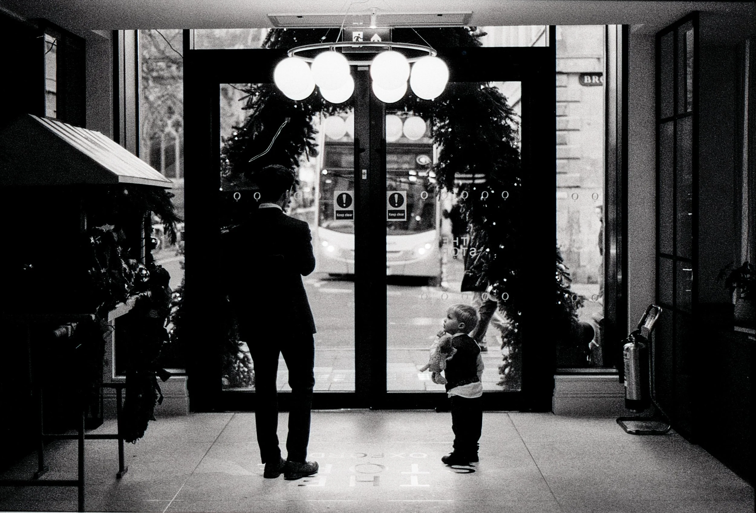 A silhouette of a man and a child facing each other inside a building near the entrance, with a bus outside, decorated with holiday wreaths and garlands.