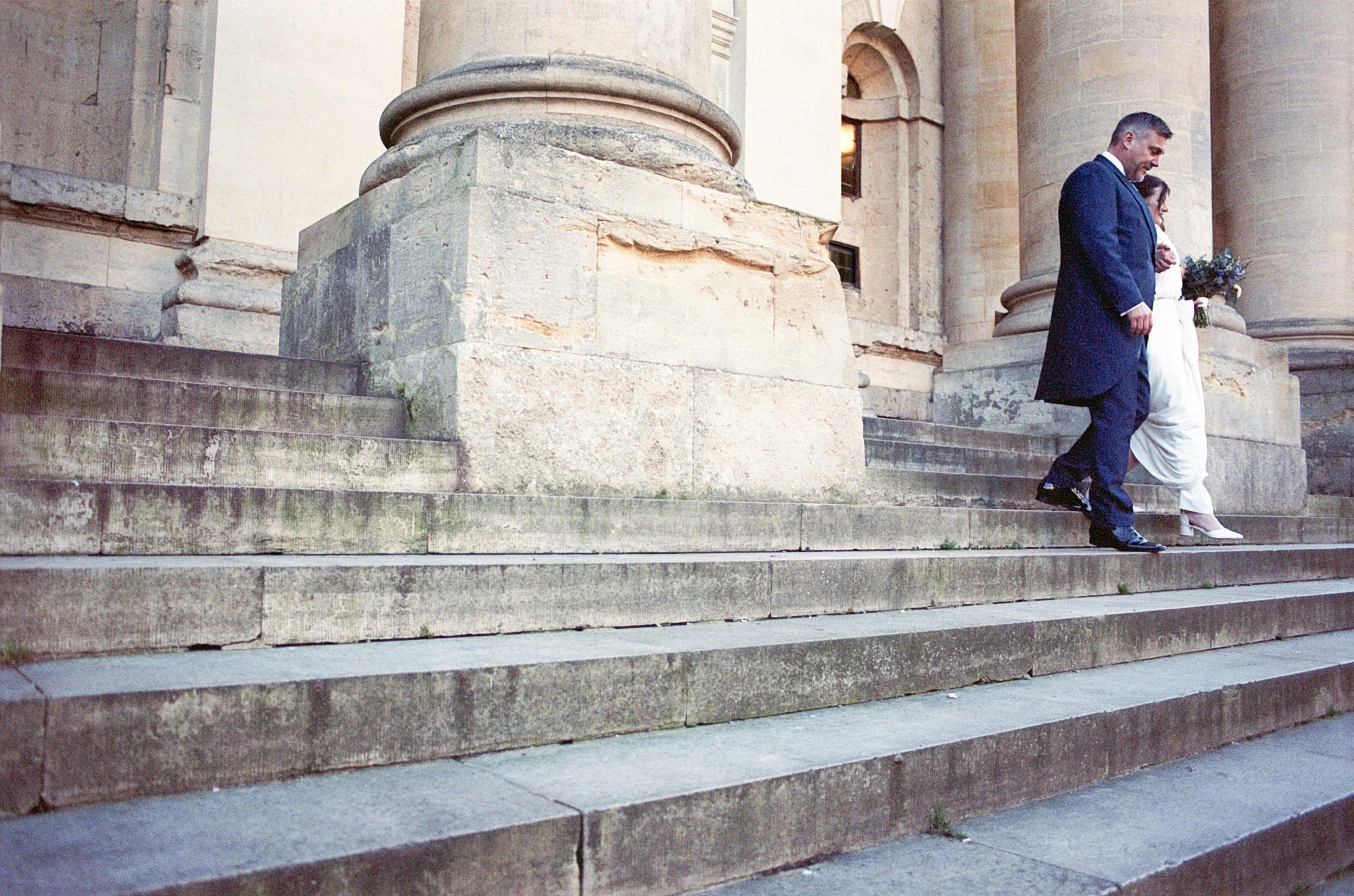 A man and a woman dressed in wedding attire walking down stone steps in front of a historic building with large columns.