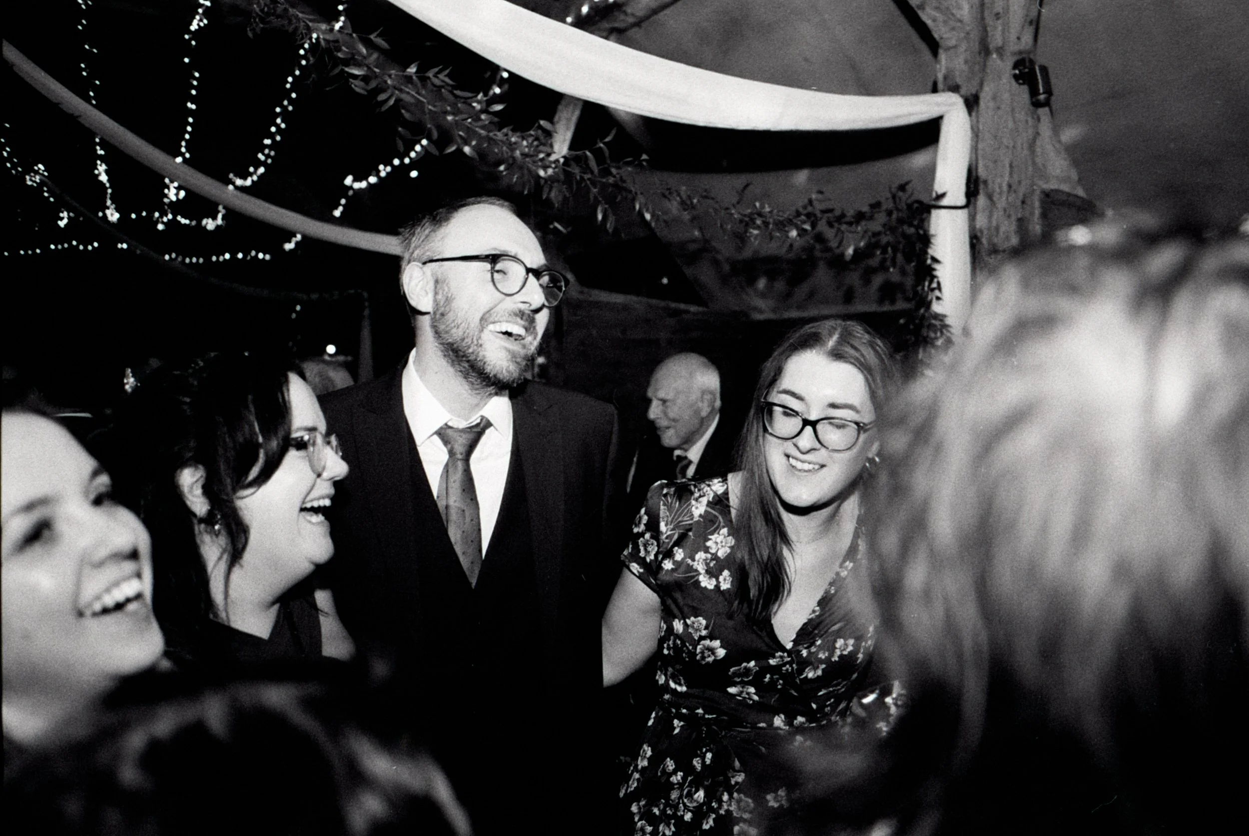 A group of people at a celebration or party, laughing and talking, with string lights and decorative drapery overhead, in a dimly lit indoor setting.