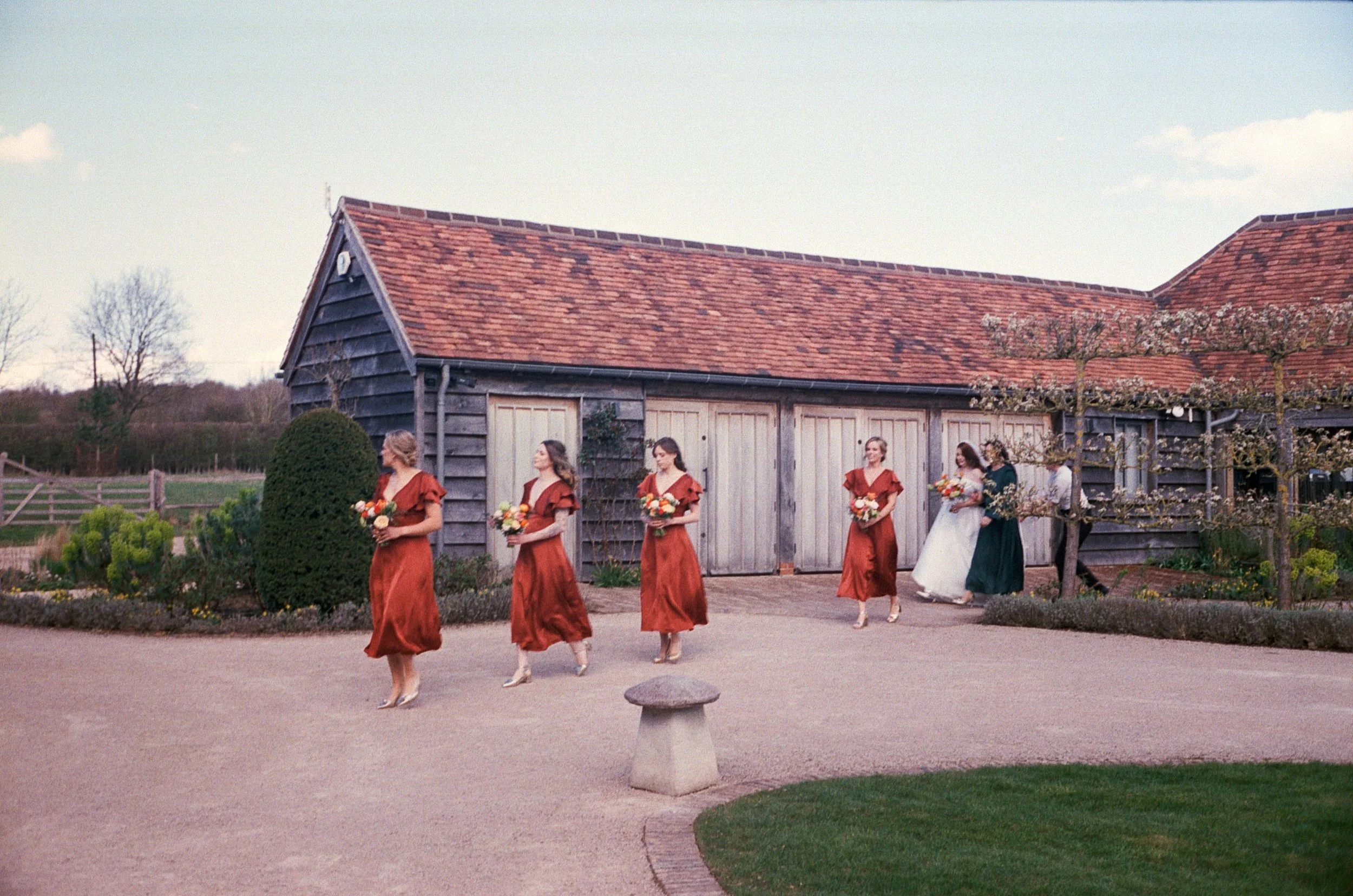 A bride and bridesmaids walking outside a rustic barn, holding bouquets of flowers, on a sunny day.