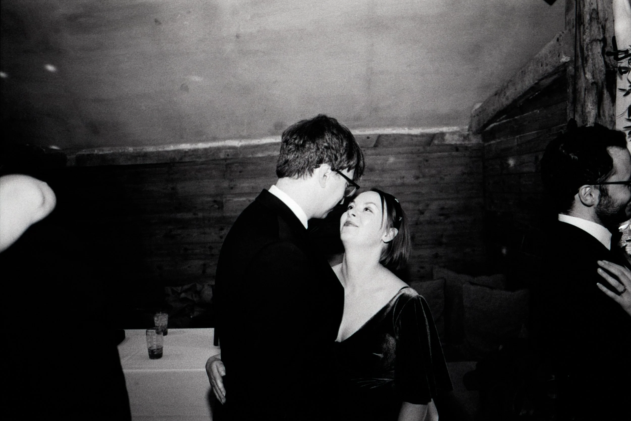 A black-and-white photograph of a couple dancing closely at a social event. The man with glasses and a suit gazes into the woman's eyes, who is wearing a dress and looking up at him. Other people are partially visible around them, with a wooden wall 
