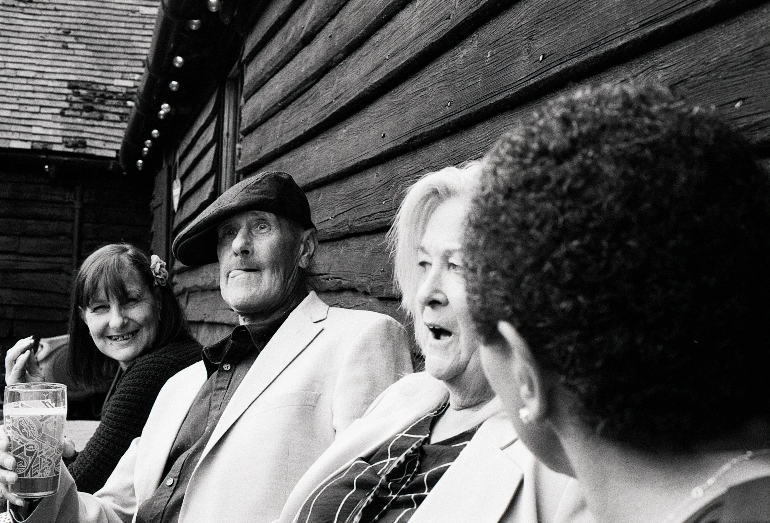 Four people sitting outdoors against a wooden wall, appearing to enjoy conversation and drinks.