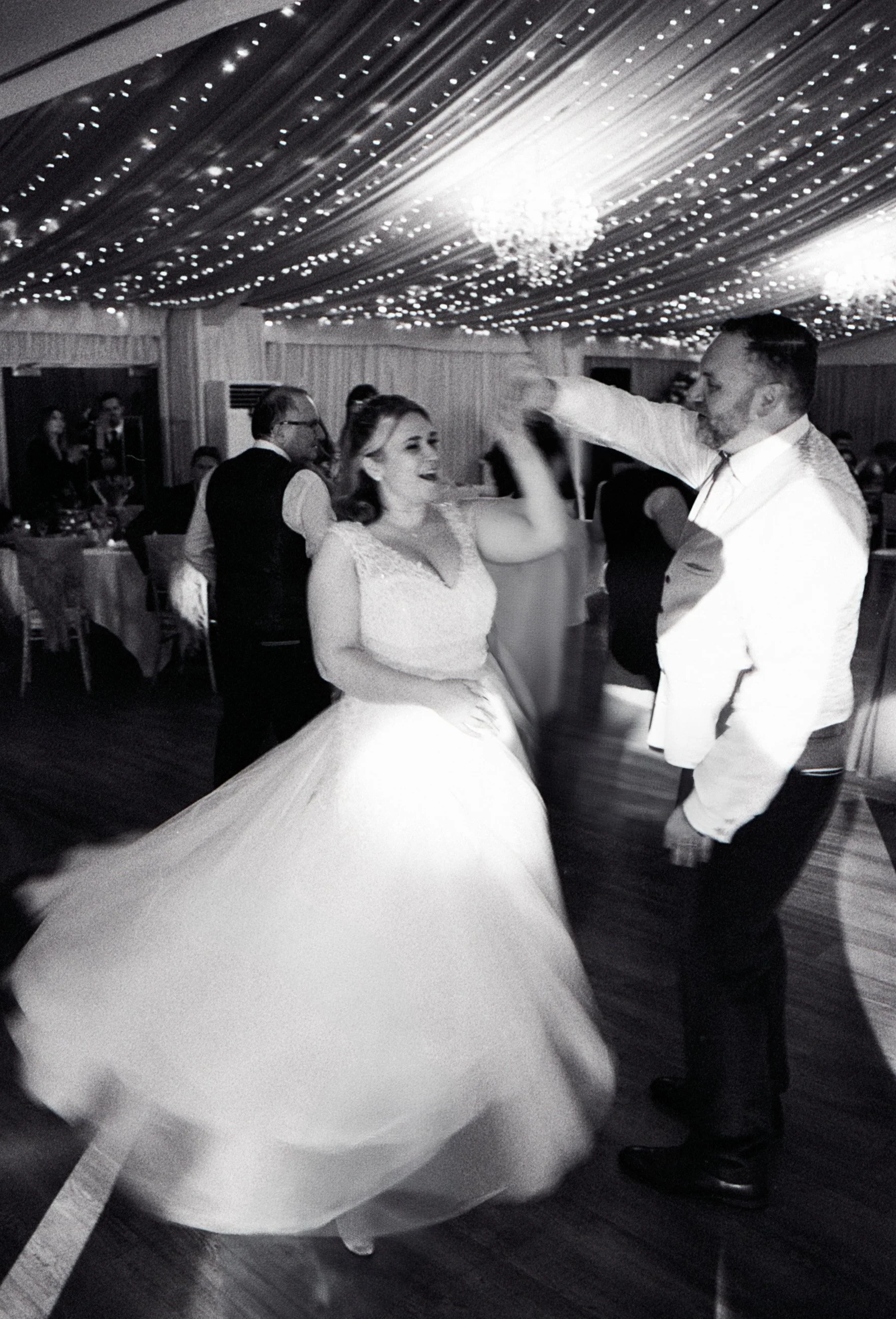 A bride and groom dancing at a wedding reception under decorated ceiling with string lights and chandeliers, with guests seated at tables in the background.