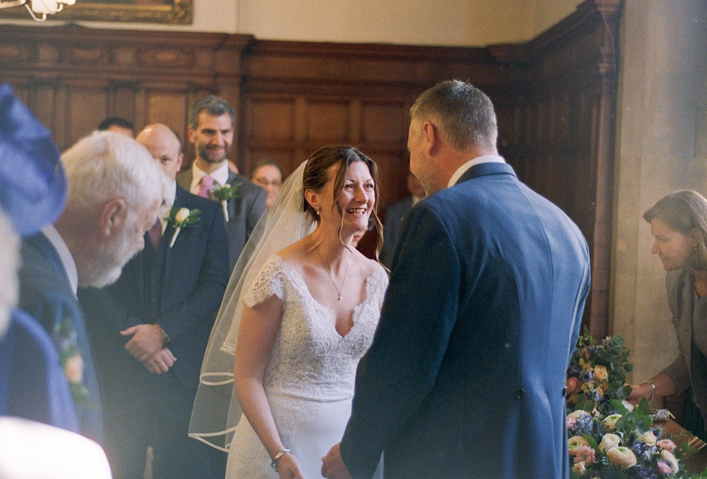 A bride and groom holding hands and smiling during their wedding ceremony indoors, surrounded by friends and family.