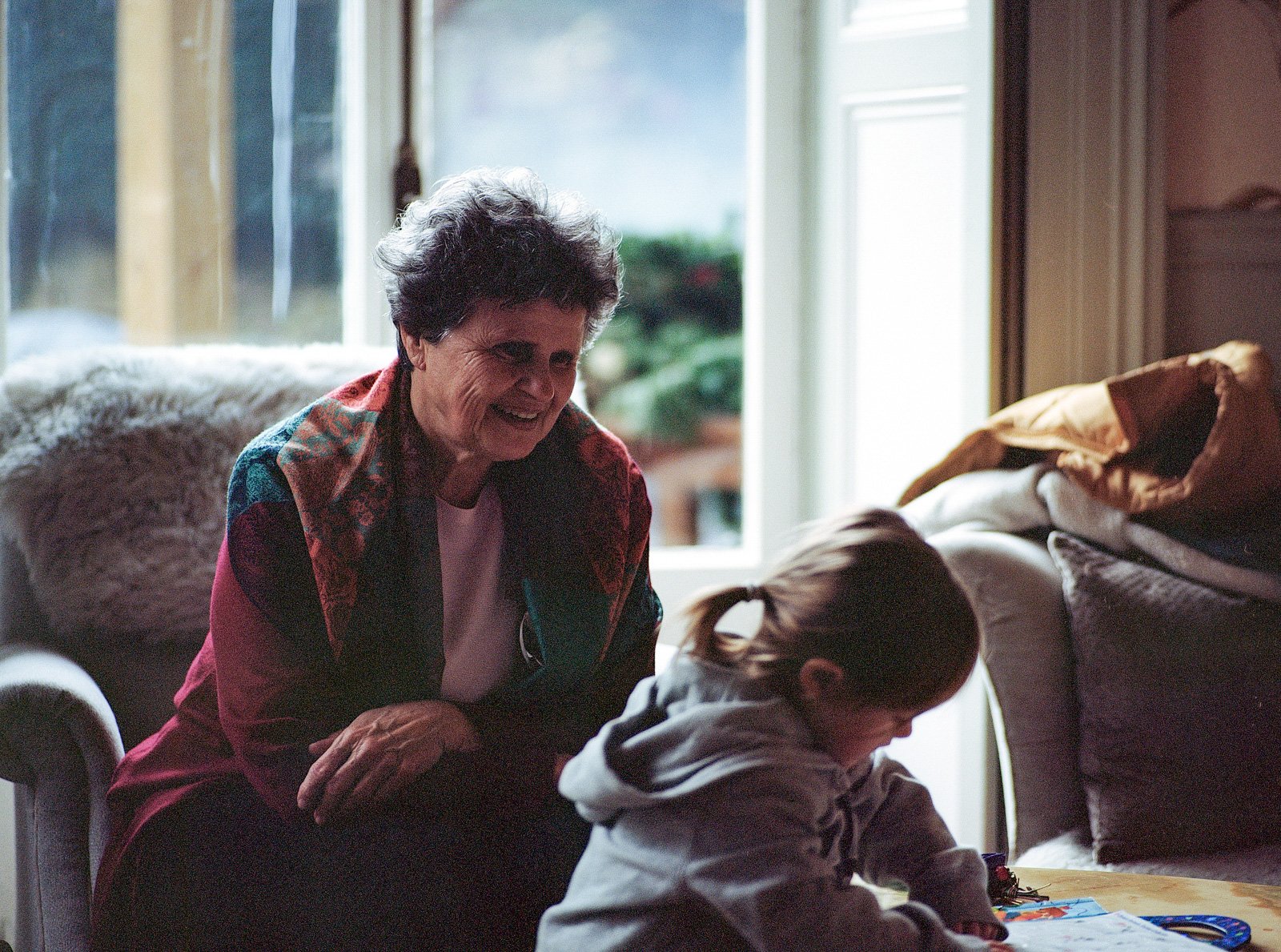 An elderly woman with curly dark hair and a colorful scarf smiling and leaning towards a young girl with light brown hair in a ponytail, wearing a gray hoodie, who is sitting at a table engaging with drawings or crafts. They are inside a cozy living 