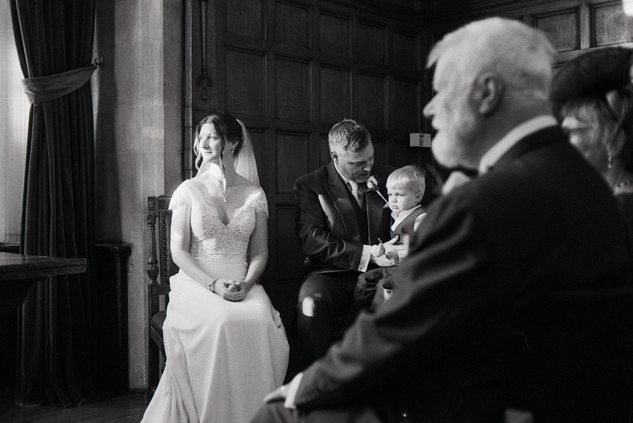 Black and white photo of a wedding ceremony with a bride in a white dress sitting on a chair, a man holding a child, and several other seated guests in a wood-paneled room.